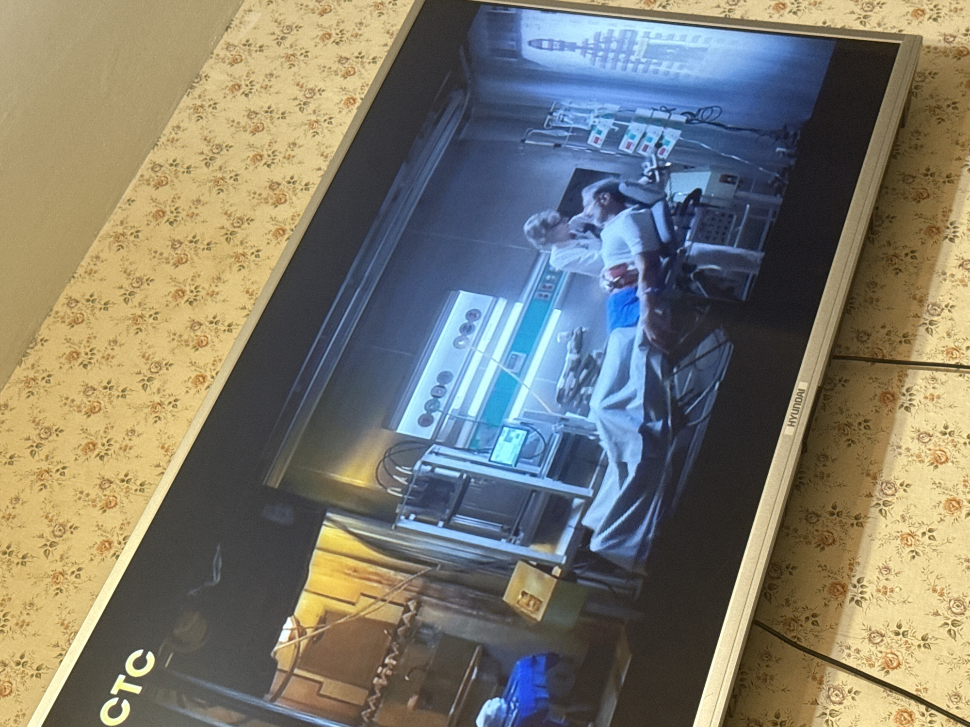 The image shows a Hyundai TV screen displaying a scene from what appears to be a medical drama or science fiction movie. The setting is a futuristic or highly advanced medical lab, with complex equipment, monitors, and fluid bags hanging overhead. Two people are visible in the scene, one wearing a white lab coat and blue scrubs, possibly a surgeon or medical professional. The environment suggests a sterile, high-tech, clinical atmosphere. The TV is placed on a surface with a floral pattern. The location in the city of Mytishchi, Russia is not possible to determine from the image.