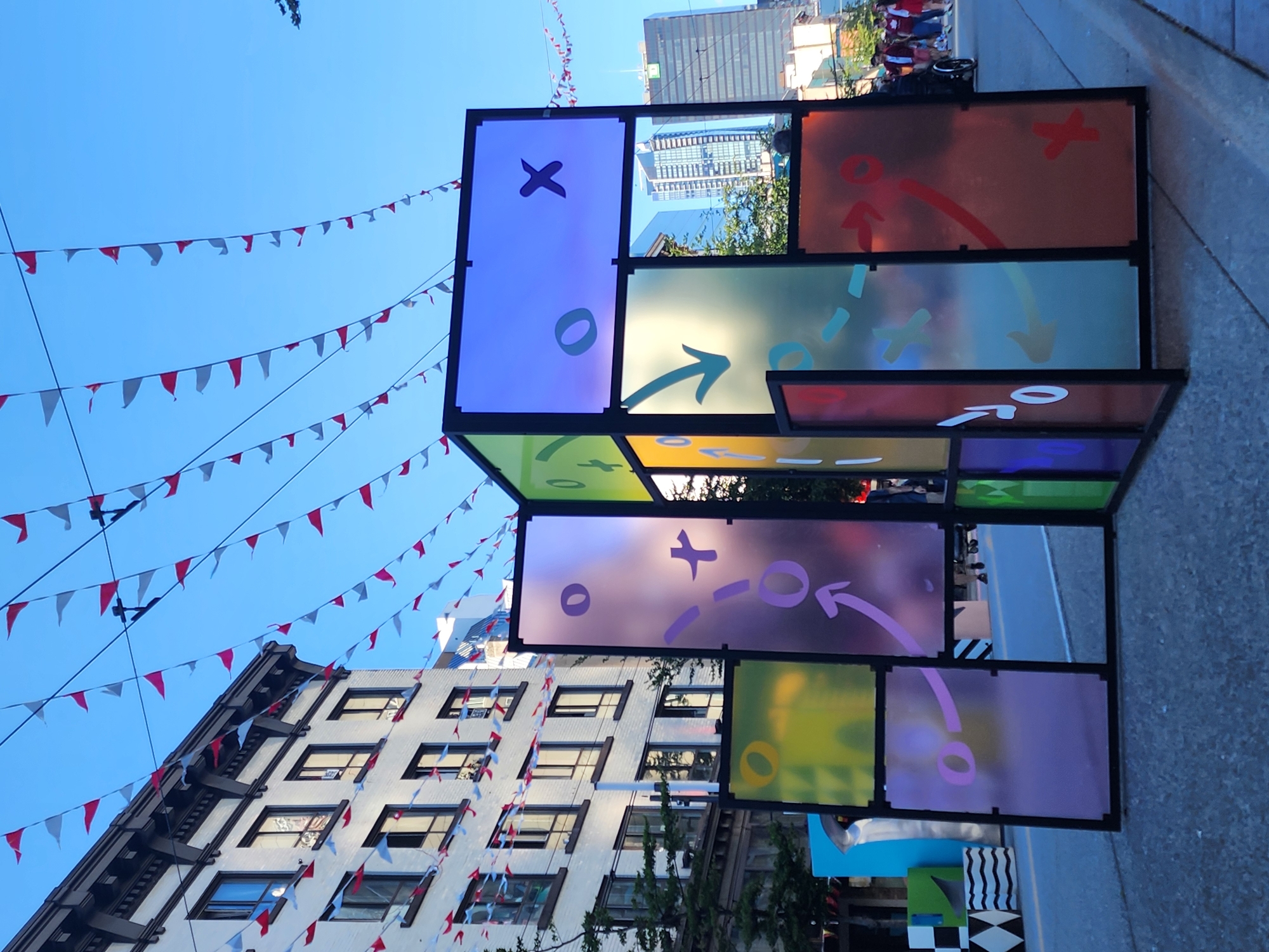 This is a medium-angle, eye-level outdoor photograph of a colorful, abstract art installation in an urban setting. The installation is made of translucent panels in various colors, including purple, green, yellow, and orange, framed by black metal. The panels are decorated with white and purple graphic elements that resemble game pieces or strategic markings, such as "X"s, "O"s, arrows, and crosses. Some panels are transparent, allowing views of the background, while others are more opaque.
Behind the art installation, a multi-story building with white brick and dark-trimmed windows is visible. Above the building, strings of red and white triangular flags are strung across the sky. In the background, modern skyscrapers rise against a clear blue sky.
On the right side of the frame, a paved street is visible, with some people walking and a person in a wheelchair. The ground in the foreground is asphalt, with concrete curbs. The overall impression is that of a public art display or an event taking place in a city street. The location is described as Unknown, Unknown.