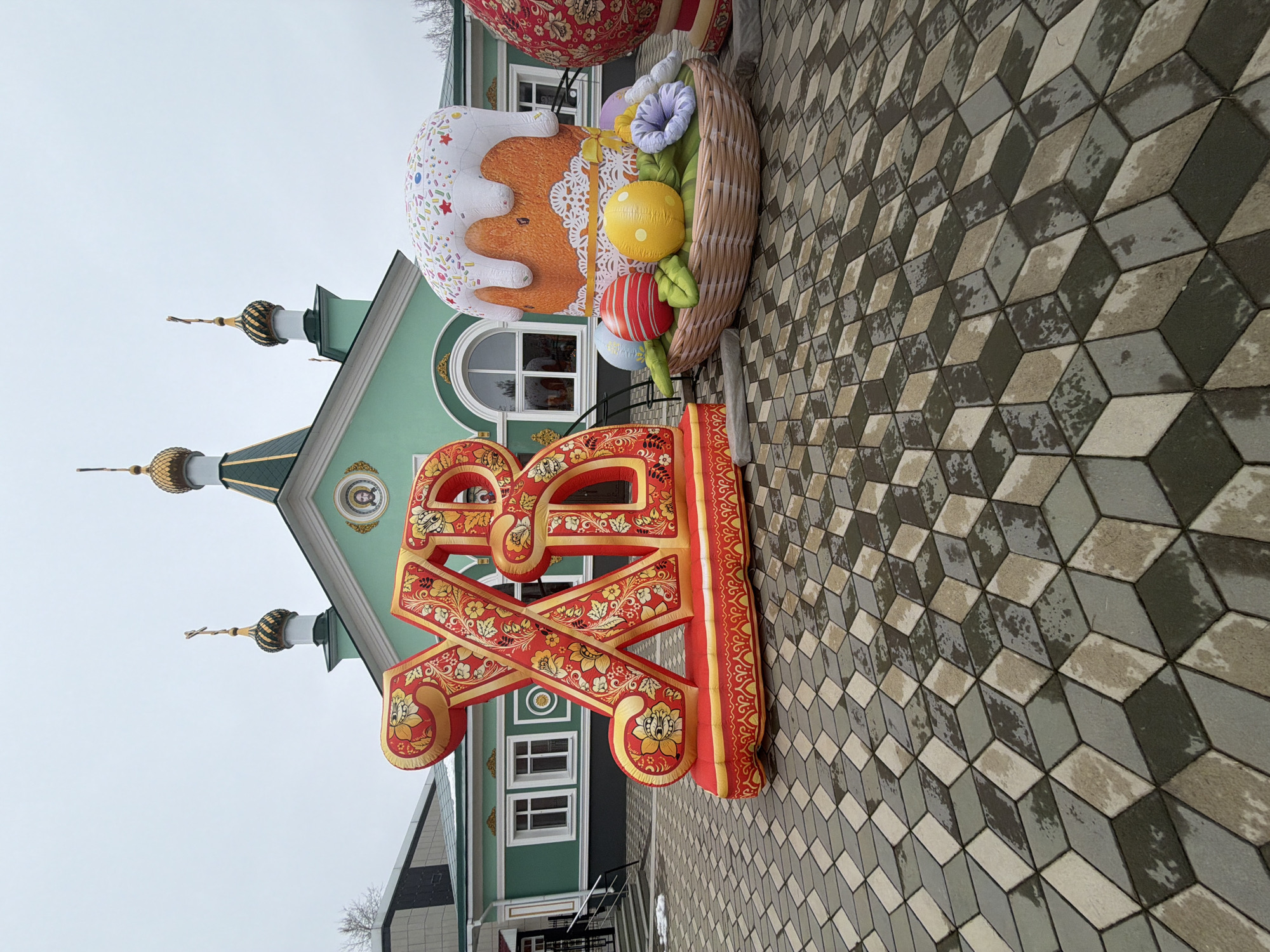 The image shows an Easter display in front of a green and white building in Chaikovsky, Russia. The display includes a large red and gold "XB" symbol, a basket of colorful eggs and flowers, and a giant Easter cake. The building has three golden onion domes with crosses. The ground is covered in gray and white patterned pavers. The sky is overcast.