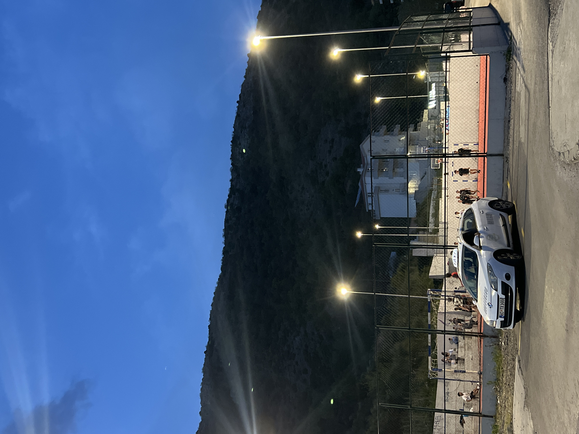 The image depicts an outdoor scene in Boreti, Montenegro, at dusk. A large mountain forms the backdrop, illuminated by streetlights. A white taxi with license plate "BD-TX-499" is parked along the right side of the frame on a paved area. A sports field, likely a soccer field, is visible behind a chain-link fence. People appear to be engaged in a game or training on the field. Buildings are visible in the background, behind the field. The sky is a gradient of blue. Streetlights line the perimeter of the field.