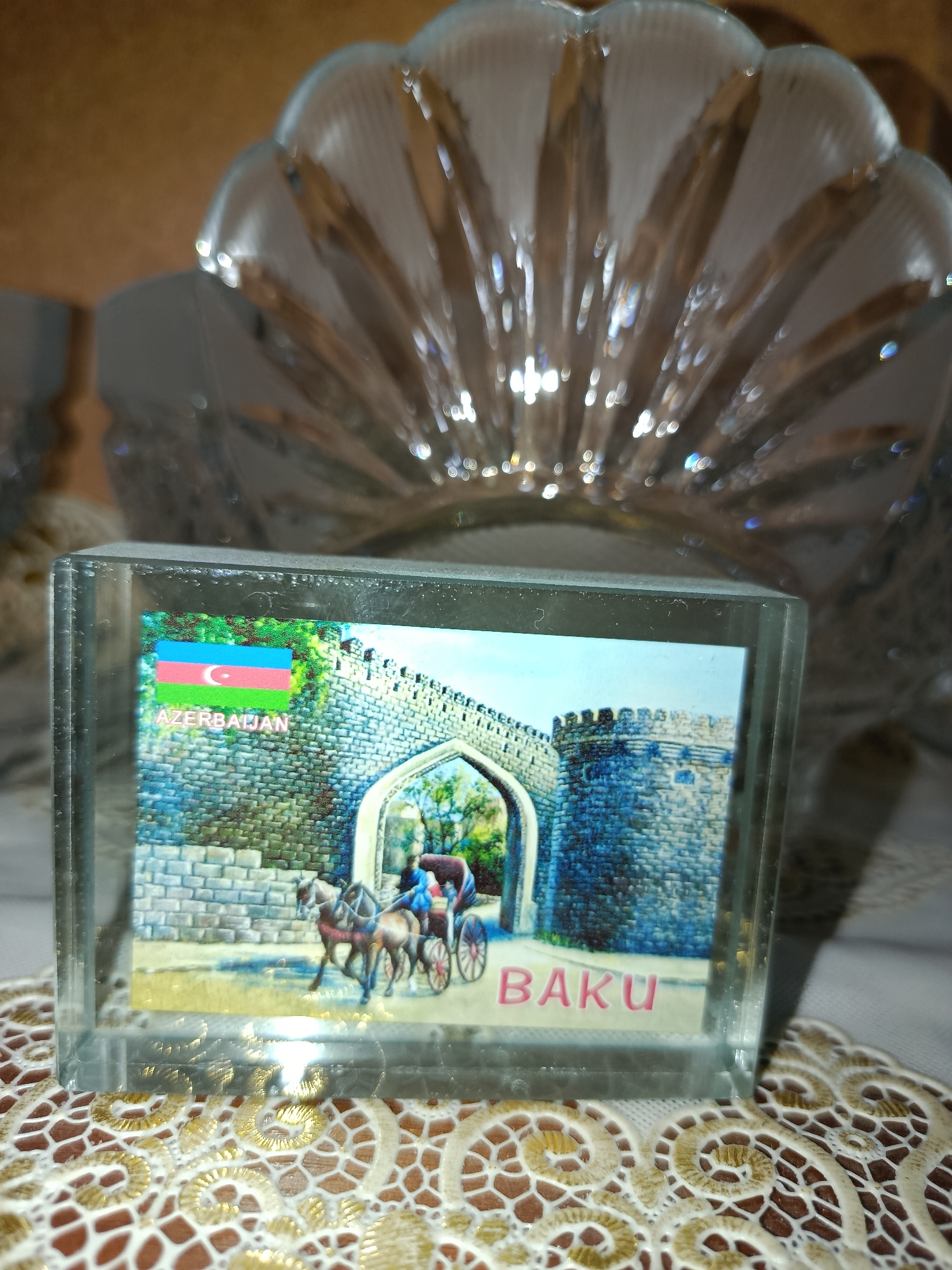 A clear glass paperweight with an image of an old city gate and a horse-drawn carriage is displayed on a beige, ornate tablecloth. The image features the flag of Azerbaijan and the word "BAKU" in pink lettering. Behind the paperweight, a large, decorative glass bowl with a fan-like rim is visible, catching the light. The background is warm and dimly lit.