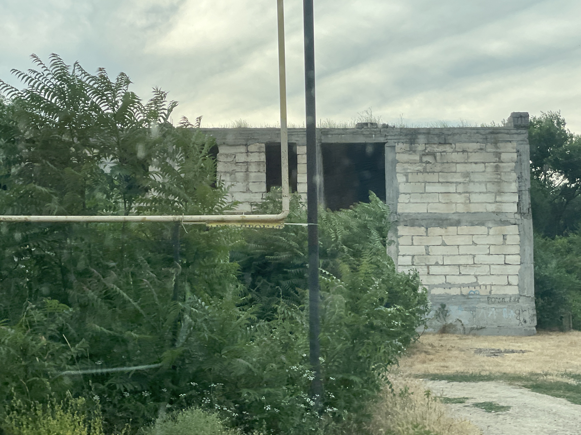 The media file depicts an unfinished or abandoned building constructed from white cinder blocks. The building is rectangular with open doorways and appears to be a single-story structure. It is partially obscured by lush green foliage, including dense bushes and trees, suggesting an overgrown or natural setting. Two vertical poles, one light-colored and one dark, are positioned in front of the building, one closer to the viewer than the other. A horizontal pipe runs across the foreground, further separating the viewer from the building.
The sky is overcast with gray clouds, indicating a potentially cloudy or dull day. The ground around the building is covered in dry grass and some scattered gravel or dirt. There is graffiti visible on the lower portion of the building's walls, with some letters and symbols. The overall impression is one of disuse and decay, with nature encroaching on the man-made structure. The time of day is unclear due to the overcast weather, but it appears to be daytime. The location is identified as Congaz, Moldova. No people or specific activities are depicted in the image.