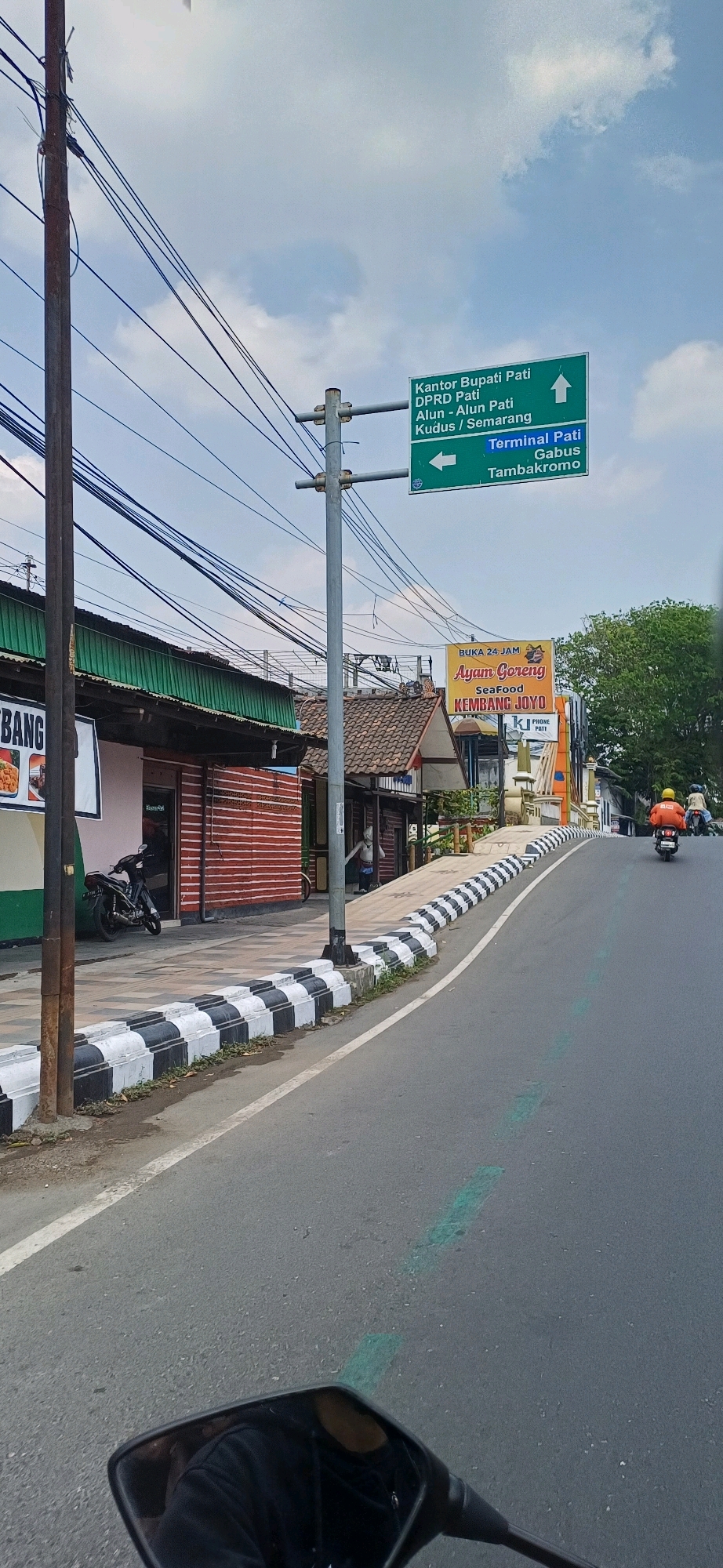 The image shows a street scene in Kecamatan Pati, Indonesia, with a clear blue sky and scattered clouds. In the foreground, a portion of a motorcycle's handlebar and a rear-view mirror are visible, suggesting the photo was taken from a moving vehicle. The road surface is asphalt, marked with a white lane divider and intermittent green markings. To the left of the road, a sidewalk with black and white checkered barriers runs alongside a row of buildings. One building appears to be a shop with red and white striped walls and a green awning, with a parked motorcycle outside. Further down the street, a yellow banner advertises "Ayam Goreng Seafood KEMBANG JOY" and "BUKA 24-JAM." A man on a motorcycle wearing a yellow helmet and an orange jacket is riding away from the camera on the right side of the road. Overhead, numerous power lines crisscross the sky, supported by utility poles. A prominent green directional signpost indicates routes to "Kantor Bupati Pati," "DPRD Pati," "Alun - Alun Pati," "Kudus / Semarang," and "Terminal Pati Gabus Tambakromo."