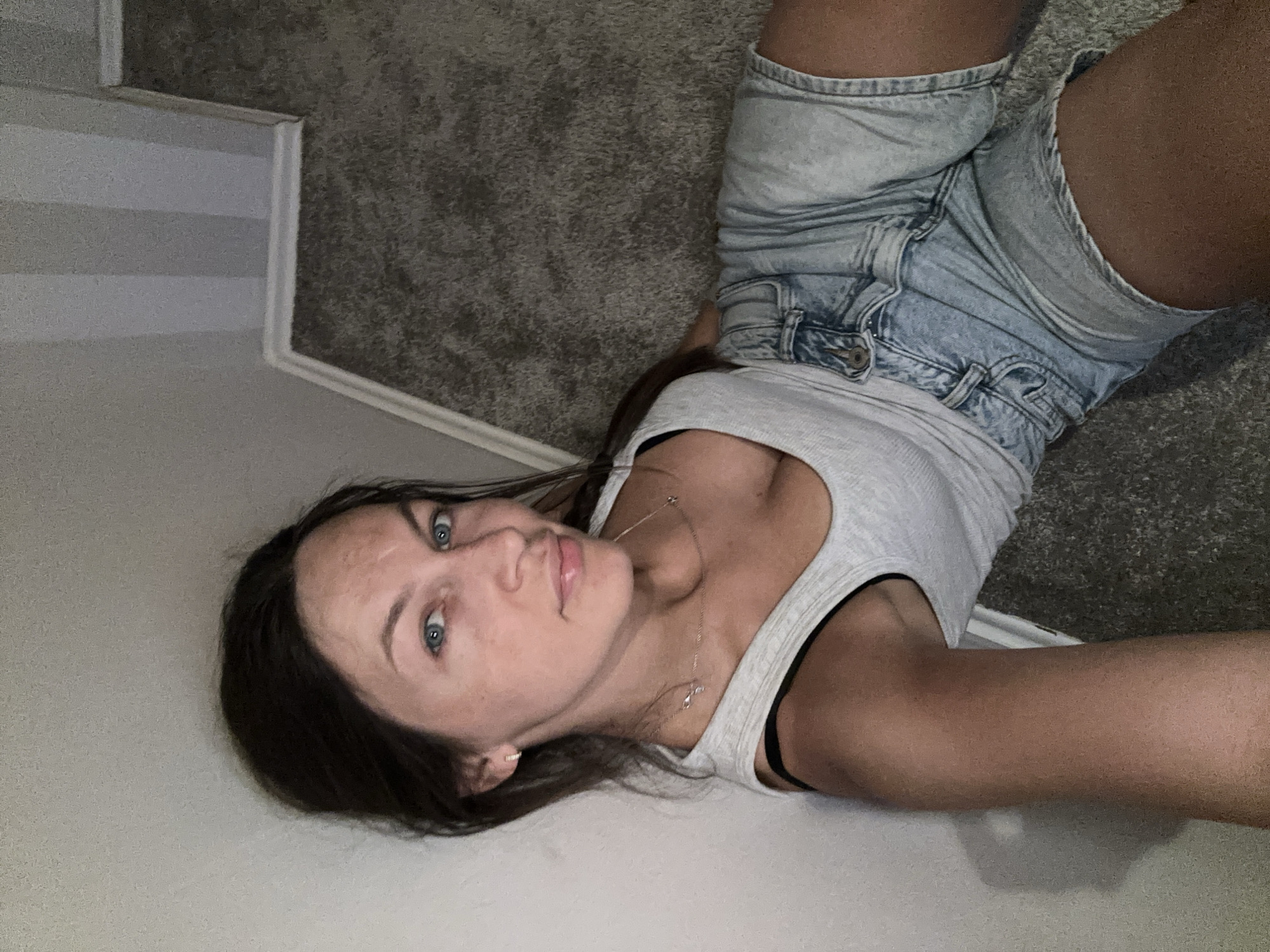 A young woman is depicted in a selfie, lying on her side on a grey, textured carpeted floor in an indoor setting in Fredenbeck, Germany. The image is oriented with her head to the bottom left and her legs extending to the top right. She is wearing a light grey tank top with black straps visible underneath, and light blue denim shorts. A delicate silver necklace with small charms is visible around her neck.
She has dark hair and striking blue eyes, and her gaze is directed straight at the camera. Her facial expression is neutral. Her left arm is extended, likely holding the camera for the selfie, while her right arm is bent, supporting her body on the floor.
The background consists of the grey carpet and a light-colored wall, with a white baseboard running along the junction of the wall and floor. A section of the wall to the left features subtle horizontal stripes of varying grey and white tones. The lighting appears to be artificial, casting some soft shadows. No visible text is present in the image.