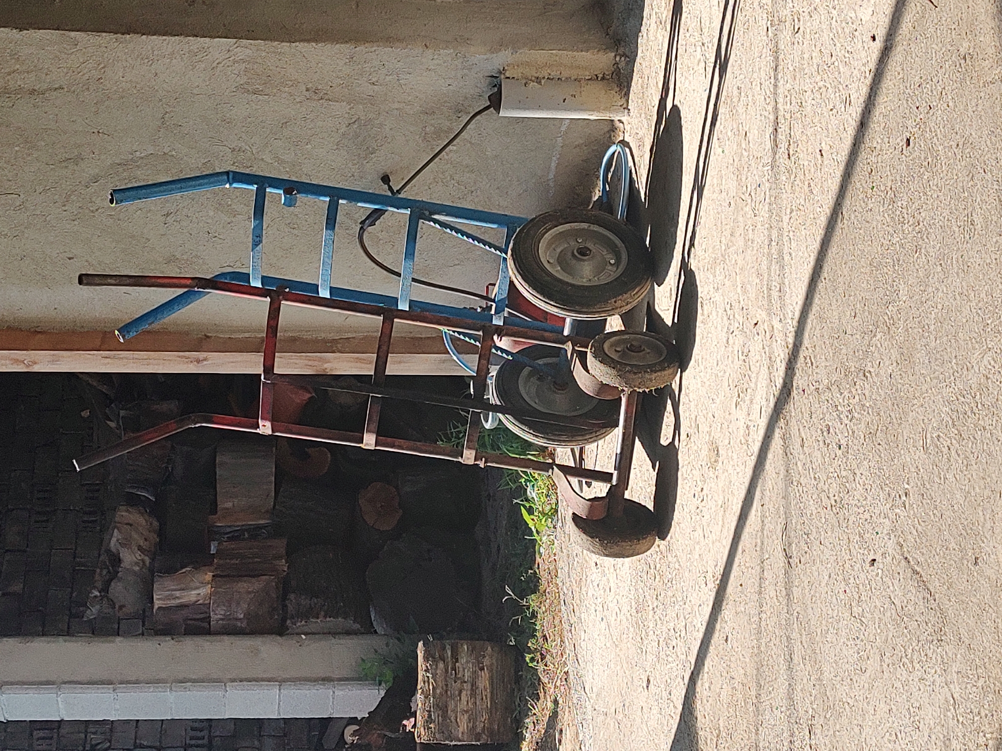 The image shows two hand trucks, one red and one blue, standing side-by-side on a dirt or gravel surface. They are positioned in front of a textured, light-colored wall, likely made of concrete or stucco, and to the left of the hand trucks is a dark, shadowy opening filled with stacked logs. A white pipe is visible next to the wall on the right. The ground is uneven and shows evidence of scattered debris and vegetation. Long shadows are cast across the ground from the hand trucks and the structures behind them, indicating sunlight from the right. The scene is devoid of any human presence. Based on the architecture and natural elements, the location appears to be an outdoor storage or work area, possibly in a rural setting.