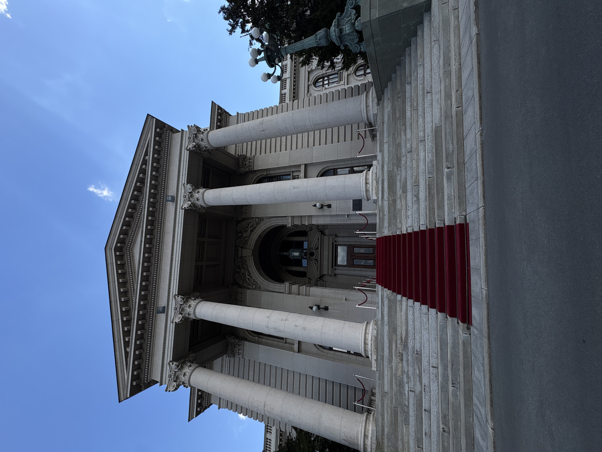 The image captures the House of the National Assembly of Serbia, located in Belgrade. The building is a grand structure with classical architectural elements, including a large portico supported by massive columns with ornate capitals. The façade is made of light-colored stone. A red carpet covers the central portion of the wide stone staircase leading up to the entrance. Ropes line the stairs on either side of the carpet. The sky is a clear blue with some light cloud coverage. A street lamp is visible at the upper right of the image.