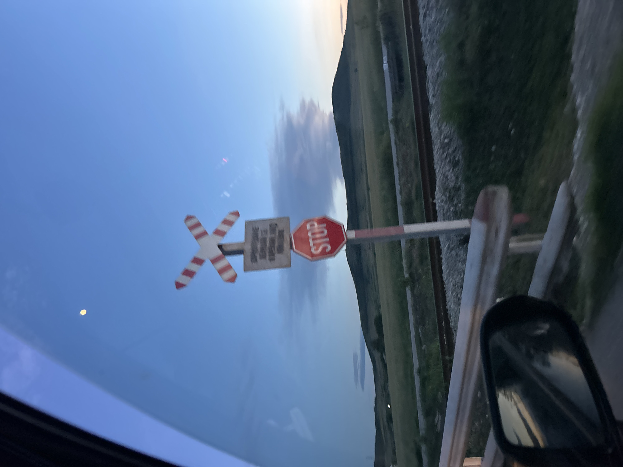 The image shows a view from inside a vehicle, likely a car. The scene is focused on a railroad crossing in Ovchepoltsi, Bulgaria. A railway crossing signal is visible, featuring a striped "X" crossing sign, a rectangular sign, and a stop sign. Railroad tracks are adjacent to the road. The sky above is blue, with a few clouds and what appears to be the moon. The vehicle's side mirror is visible in the foreground.