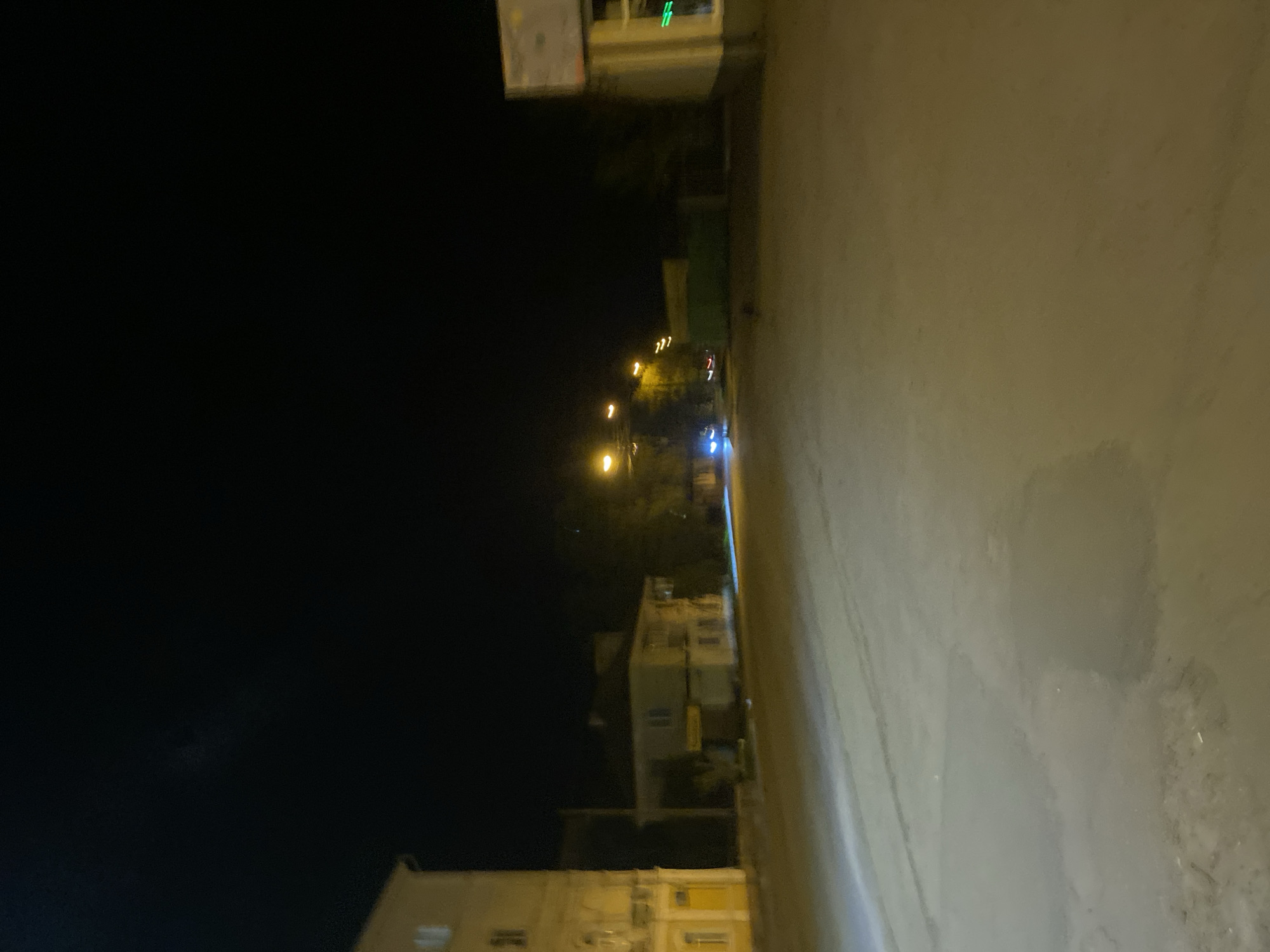 The image depicts a night scene in what appears to be a residential area of Poltava, Ukraine. The frame is divided between a dark, black sky on the left and a blurred street on the right. Streetlights are visible in the distance, casting a yellow glow, with hints of a building on the horizon. There are buildings with lit windows on the left side of the image. The street surface is wet, possibly from recent rain. The perspective is somewhat skewed, possibly from a handheld camera.