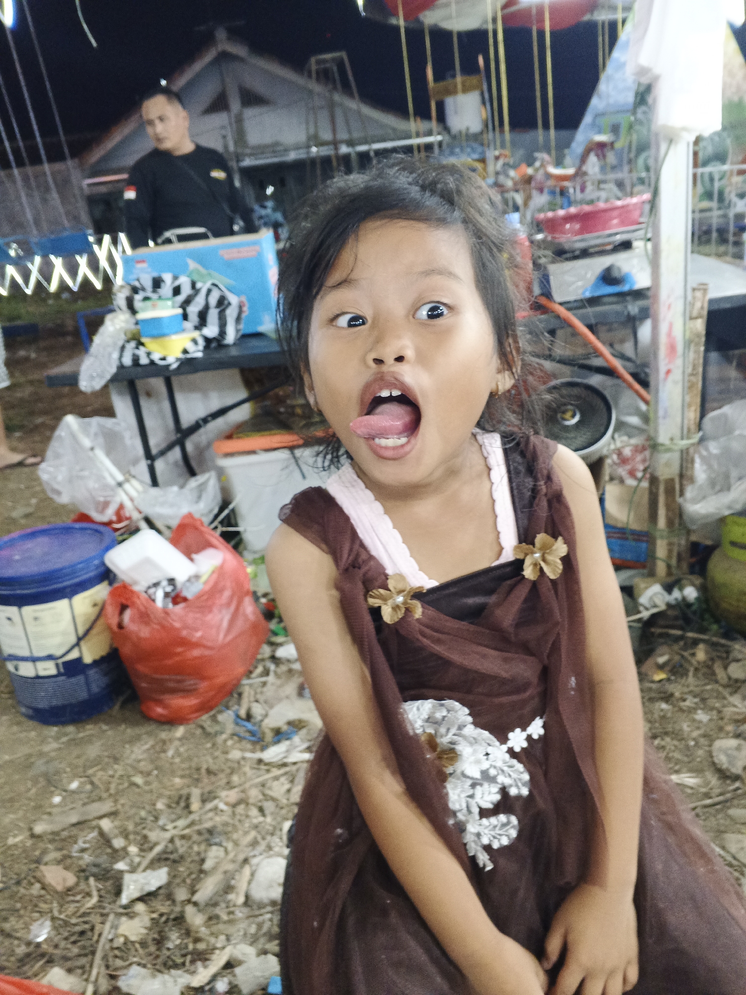 The image shows a young Indonesian girl with her tongue sticking out, making a funny face. She is wearing a brown dress with floral embellishments. In the background, there are carnival rides and a man in a black shirt. The scene appears to be at a local fair or carnival. The ground is littered with debris, suggesting an outdoor, possibly temporary setup. The image was taken at night, as indicated by the artificial lighting. This appears to be a candid shot, capturing a moment of playful interaction.