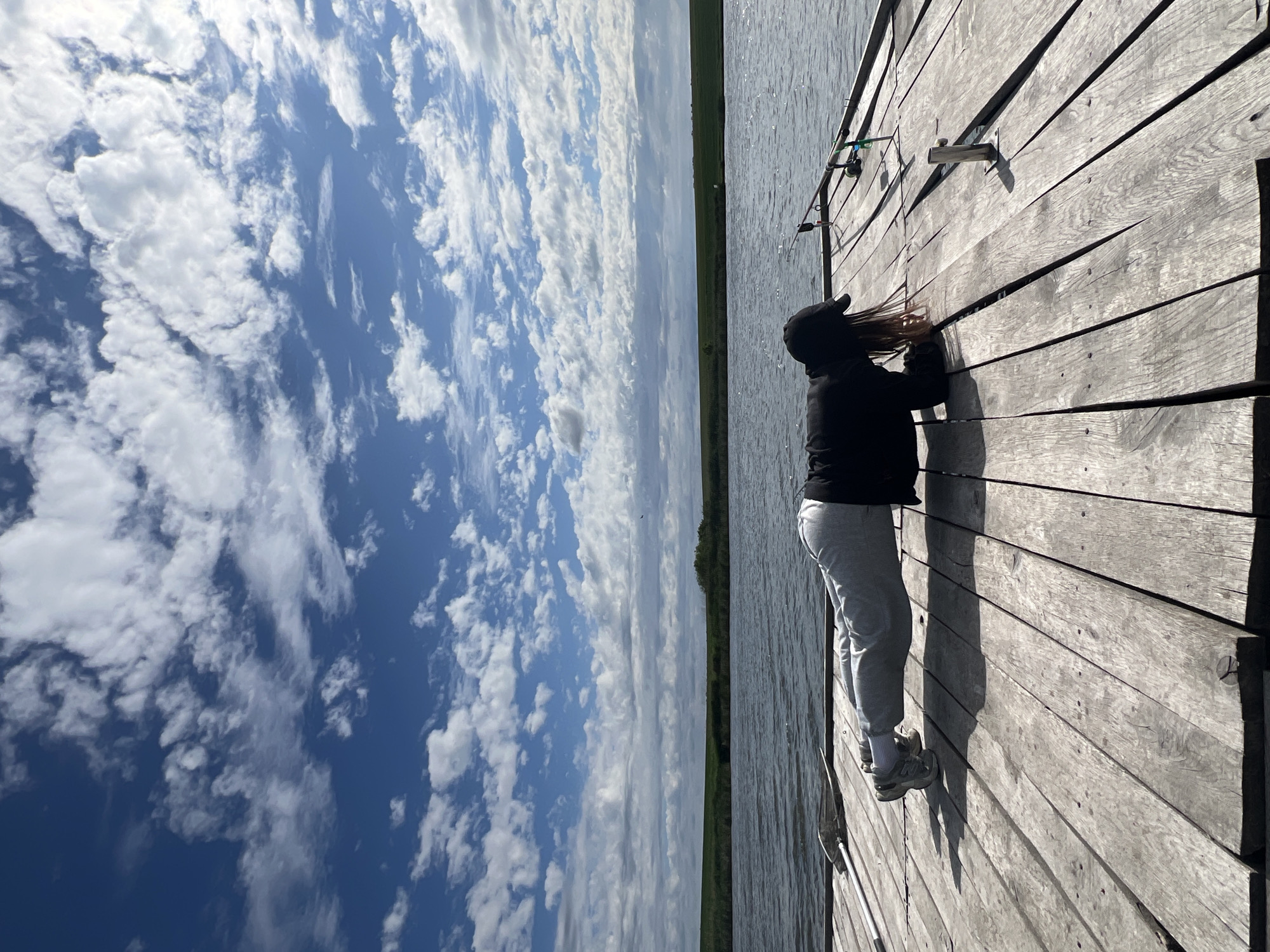 A woman stands on a wooden pier next to a body of water on a sunny day. The pier is constructed of gray, weathered wooden planks. The woman is wearing a black jacket, gray sweatpants, and a black baseball cap. She appears to be adjusting something on the pier near two fishing rods. The water is calm. The sky is mostly blue with white clouds. A green strip of land is visible across the water. The location is Kopychyntsi, Ukraine.