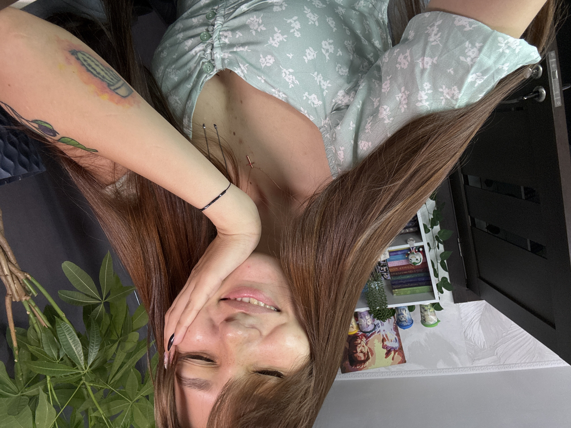 The image shows a young woman with long brown hair, wearing a light blue dress with a floral pattern, indoors. She is resting her head on her hand and smiling. Behind her, shelves are visible with books and decorative items, including what appears to be a poster of Iron Man. A large green plant is also present in the background. The environment suggests a home or personal space. There is no information available in the image to determine its specific location within Novokuznetsk, Russia.