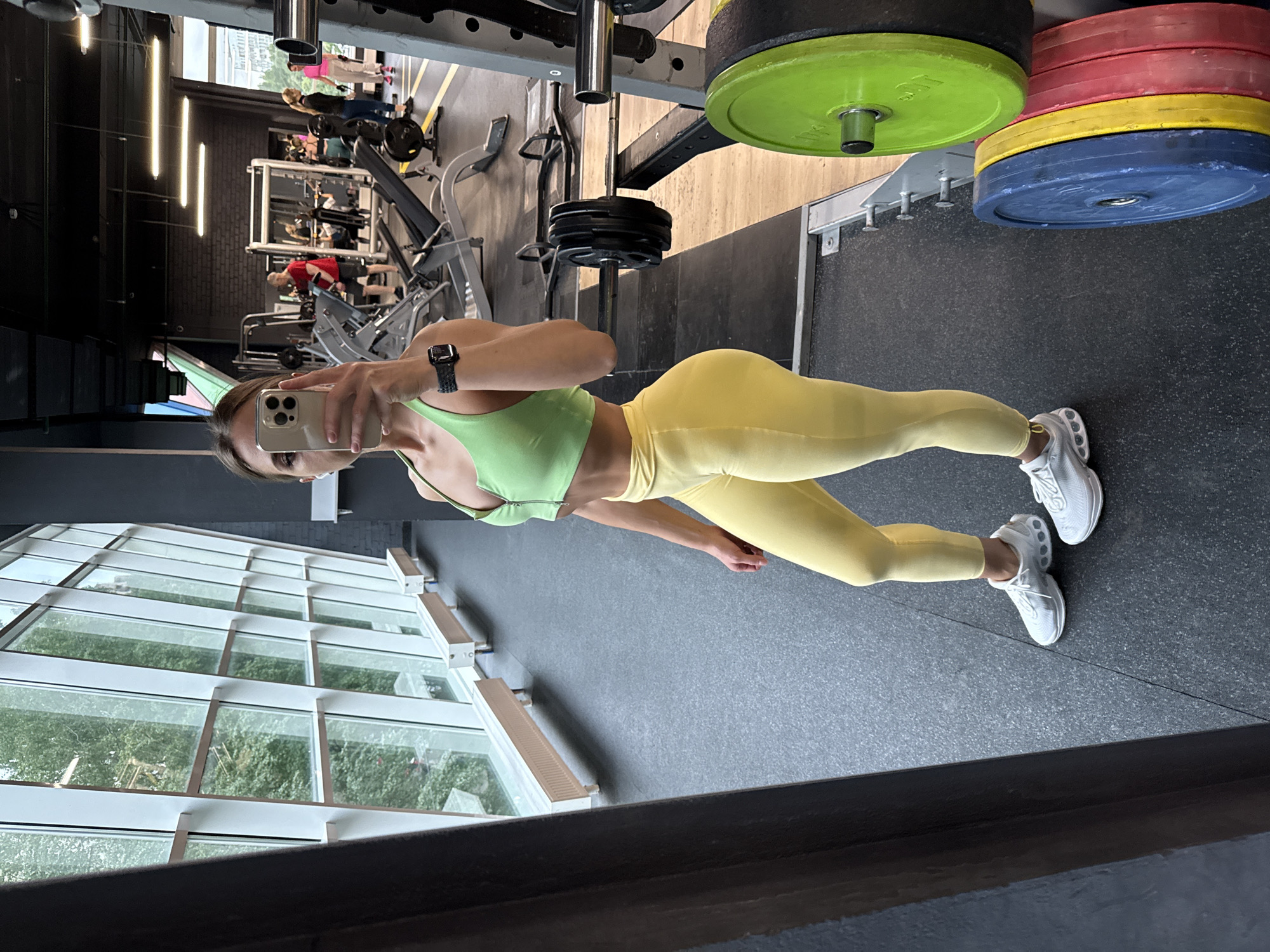 The image is taken in a gym, possibly in Saint Petersburg, Russia, given the architecture seen through the large windows. A woman wearing a light green sports bra and yellow leggings is holding a smartphone to take a selfie. She is standing in front of a mirror. Gym equipment such as weights, weightlifting machines, and other gym-goers are visible in the background. The floor is a dark gray, and there are large windows that reflect greenery outside.