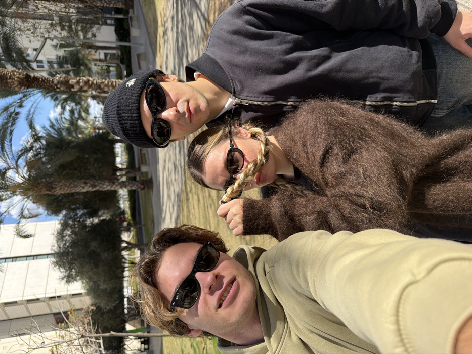 Three individuals are depicted outdoors in a modern urban park or green space in Tel-Aviv, Israel, under a clear blue sky. The background features light-colored, multi-story buildings with grid-patterned windows and numerous tall palm trees, consistent with areas such as the Tel Aviv University campus or contemporary neighborhoods.
The subject on the left, a male with light brown hair, wears a light-colored hoodie and black Wayfarer-style sunglasses. He is holding the camera to capture a selfie. The central subject, a female with blonde hair styled in two braids, wears a dark brown fuzzy sweater and small oval black sunglasses; she holds one of her braids across her face. The subject on the right, a male, wears a black beanie with a white Stüssy logo, dark oval sunglasses, a black bomber jacket, and light blue jeans. All three subjects are oriented towards the camera.