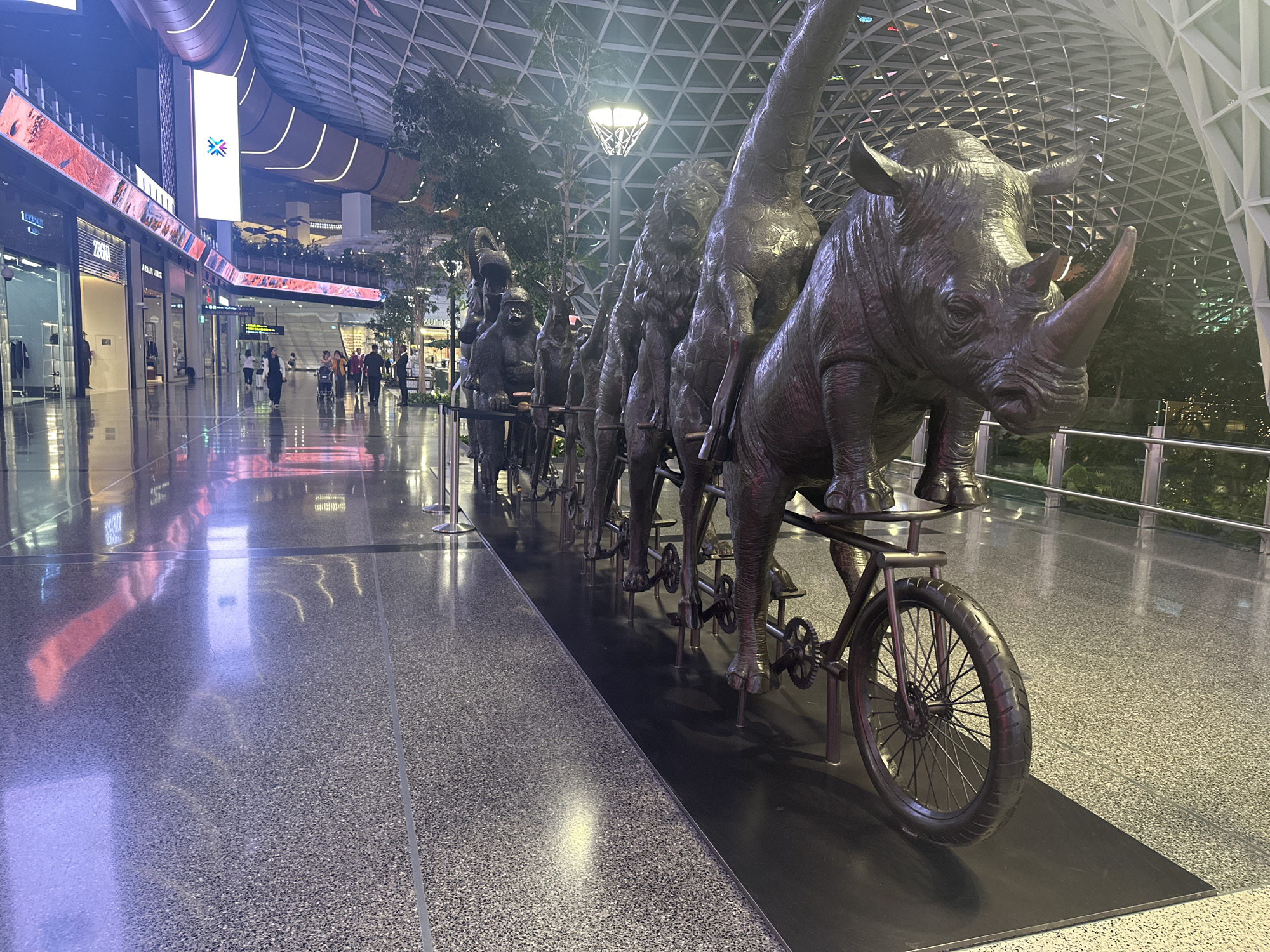 The image depicts a large bronze sculpture of various animals, including a rhinoceros, giraffe, lion, and others, depicted riding unicycles in a line. The sculpture is situated in the Hamad International Airport in Doha, Qatar, which is characterized by its modern architecture, high ceilings with geodesic dome-like structures, and extensive use of glass and reflective surfaces. The floor is polished and shows reflections of the surrounding lights and the sculpture itself. In the background, shops and escalators are visible, along with a few people walking and pushing luggage. The lighting is a mix of artificial light from the ceiling and storefronts, creating a well-lit, interior environment.