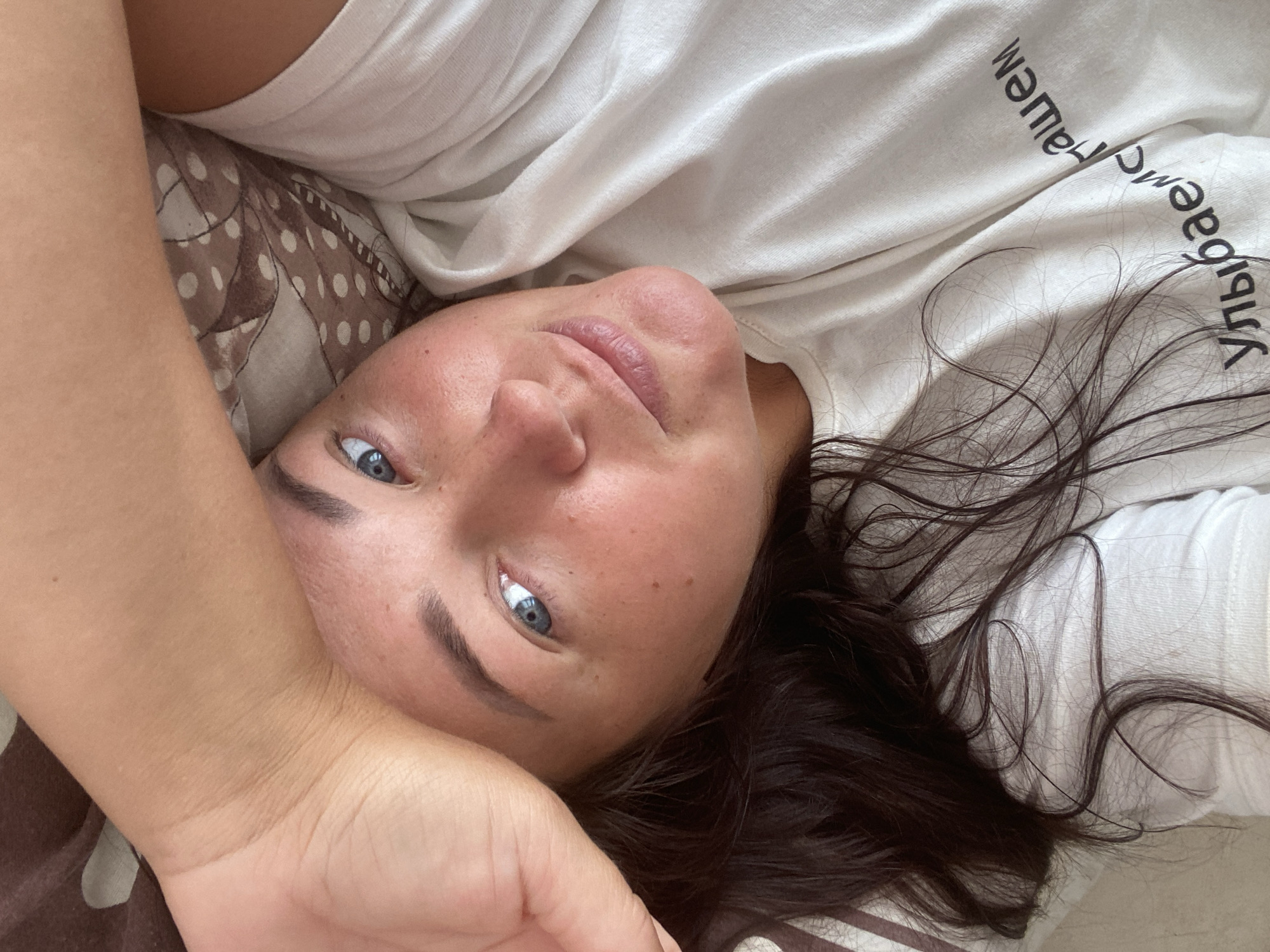 This is a close-up, upside-down selfie of a person lying down. The subject has fair skin, blue eyes, and dark brown hair. They are wearing a white t-shirt with black text printed on it. The text is in a foreign language, and appears to be in a curved pattern. The subject is lying on a bed with patterned pillowcases. The scene appears to be indoors, possibly in a bedroom. The lighting suggests it could be daytime, but no specific time can be determined. The overall mood conveyed is relaxed and casual. The visible text reads "yvibogaemc-ralllem" and is oriented in a curved manner. The setting is implied to be Sochi, Russia.