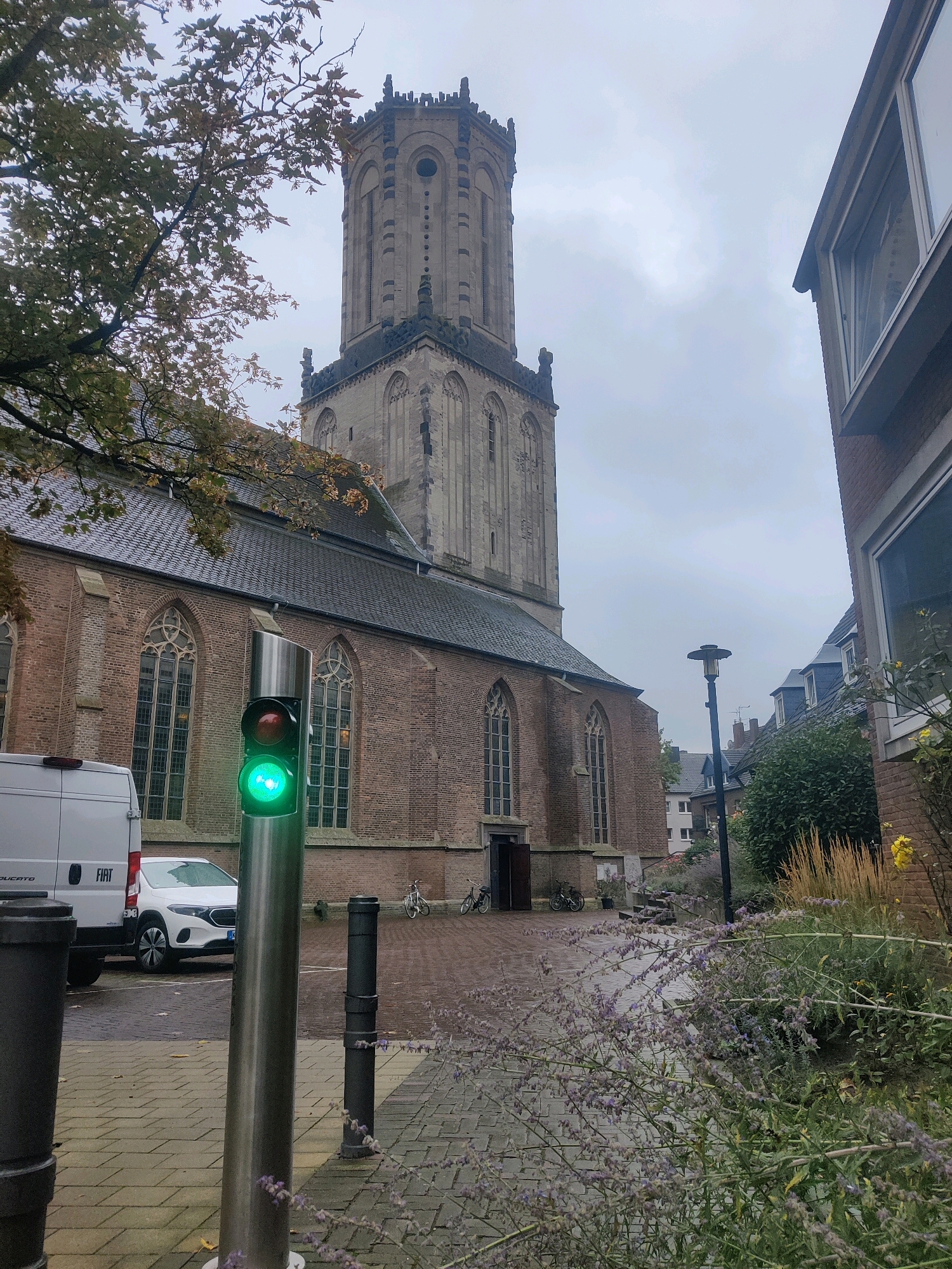 The image shows a street scene in Emmerich on the Rhine, Germany, with a tall brick church and its tower dominating the background. In the foreground, a traffic light displays a green signal. To the left, the side of a white van is visible, and a white electric car is parked. The street is paved with interlocking bricks, and the ground appears damp, suggesting recent rain. To the right, a modern building with large windows is partially visible, and beyond it, other residential buildings can be seen. Vegetation, including flowering plants and tall grasses, lines the edge of the street, with some autumnal foliage on trees. Bicycles are parked near the entrance of the church. The sky is overcast.