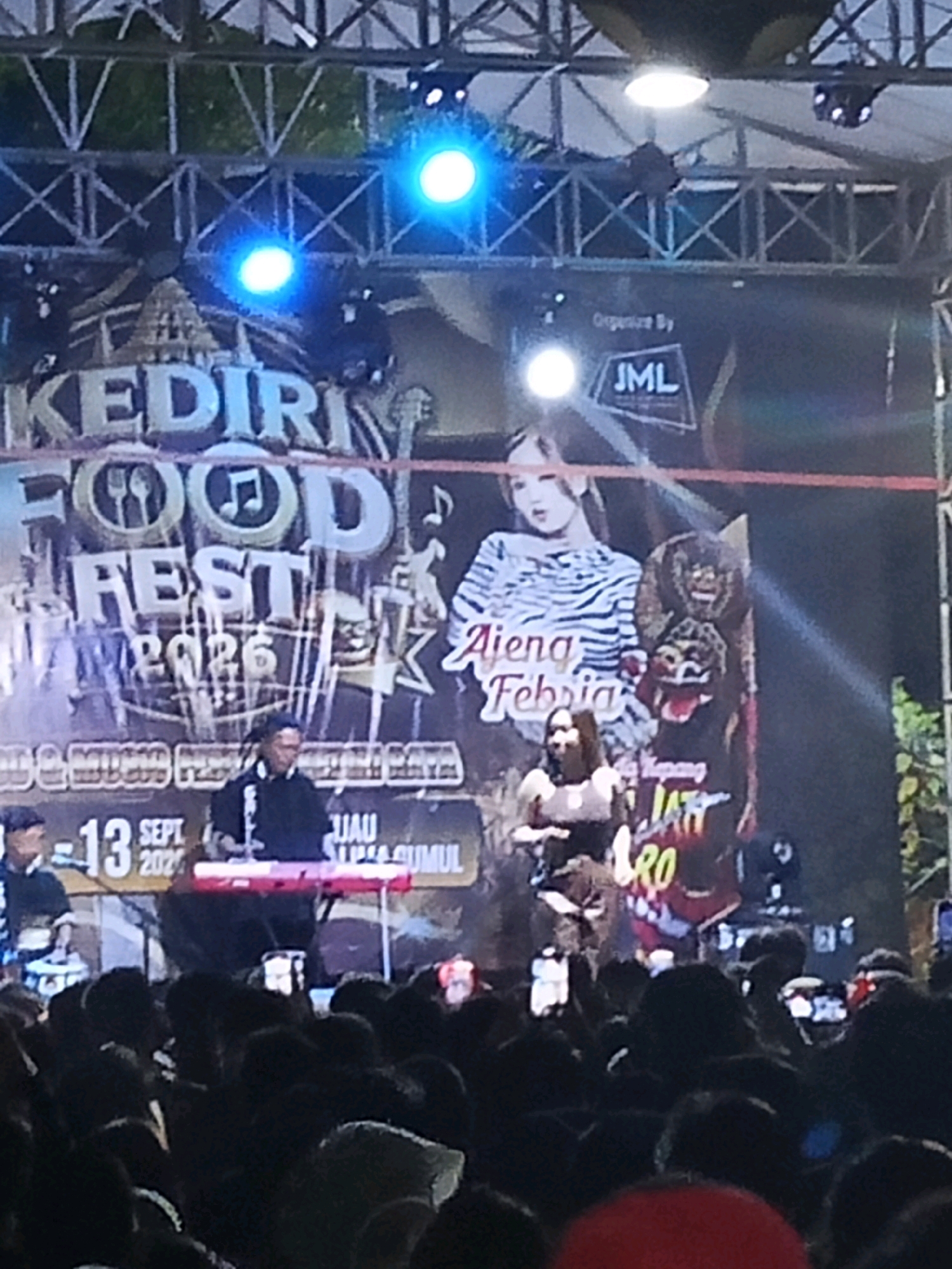 A nighttime outdoor event is captured in this image, likely a music festival or concert, as indicated by the stage, lighting rig, and the presence of a crowd. The event appears to be a "KEDIRI FOOD FEST 2023" or similar, with banners featuring the text "KEDIRI FOOD FEST" and "2023" prominently displayed on the stage backdrop. A graphic on the backdrop shows "Ajeng Febria" with a tiger-striped pattern, suggesting she is a performer.
Several individuals are visible on stage. A person in a black shirt is seated at a red keyboard or musical instrument. To their left, another person is partially visible, also appearing to be a musician. A female performer in a dark top is standing and appears to be engaging with the audience. Behind the performers, there are large graphic elements on the backdrop, including what looks like traditional masks or puppets and text in Indonesian, such as "JAU SUMUL."
The stage is illuminated by bright blue spotlights from above. A metal truss structure supports the lighting and banners. Below the stage, a dense crowd of people is visible, many holding up their phones, presumably to record or photograph the event. The overall atmosphere suggests a lively and engaging public gathering. The location is identified as Ngasem, Indonesia, based on the text on the banner and general context of such events.