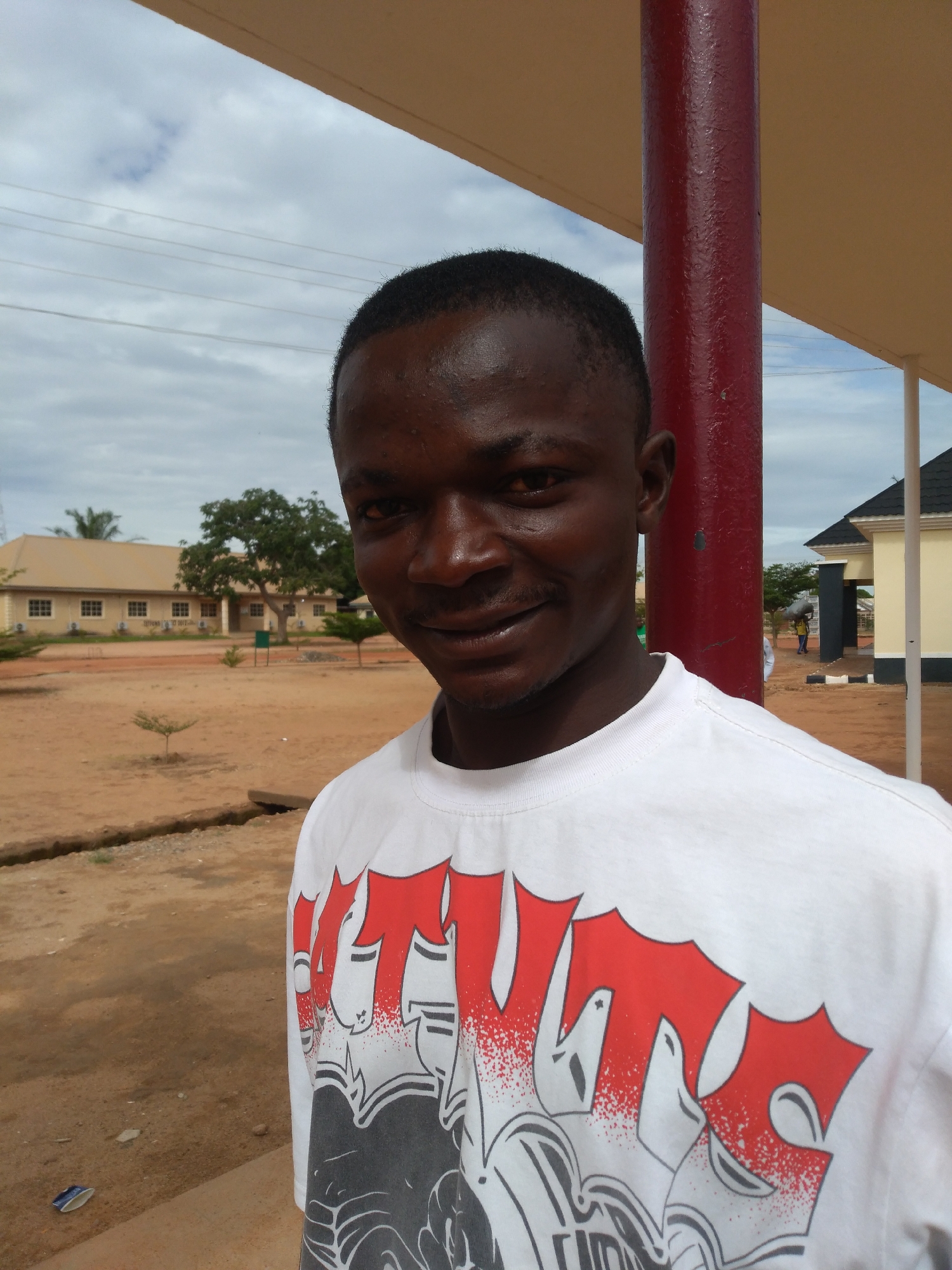 A young, dark-skinned male is prominently positioned in the foreground, slightly left of center, smiling faintly and gazing directly at the viewer. He has short, dark hair and is wearing a white long-sleeved t-shirt with a large, stylized graphic design in red, black, and white, featuring bold shapes and distressed textures.
To the subject's right, a thick, dark red cylindrical pillar stands vertically, partially obscuring the background. Above, a light brown/beige roof or awning structure is visible. The ground in the mid-ground and foreground is composed of reddish-brown earth, characteristic of laterite soil, with a visible shallow ditch or drain. Scattered small saplings and a larger tree with green foliage are present in the landscape.
In the background, to the left, a long, single-story building with a light brown exterior, numerous windows, and white external air conditioning units is visible. To the far right, another building with a yellow/cream facade and a dark, sloped roof can be seen. Faint figures of people are discernible in the distance near the building on the right, one appearing to carry an item on their head. Overhead, the sky is partly cloudy with patches of grey. Utility lines stretch horizontally across the upper left portion of the frame. The scene depicts an outdoor campus or institutional area in the city of Jos, Nigeria.