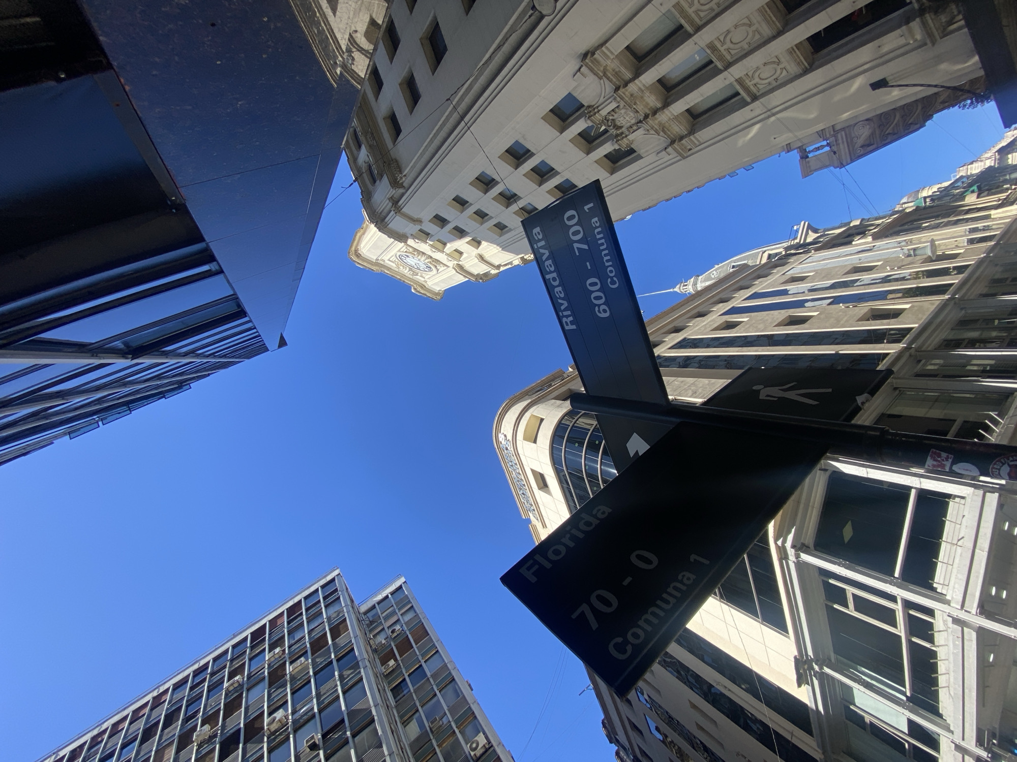 The image presents a low-angle view looking straight up at the intersection of two streets in Buenos Aires, Argentina, under a clear blue sky. Tall buildings of varying architectural styles frame the shot, creating a canyon-like effect.
In the foreground, a black street sign prominently displays street names and address ranges. One arm reads "Rivadavia 600 - 700 Comuna 1," and the other reads "Florida 70 - 0 Comuna 1." A white pedestrian symbol is also visible on the sign. The strong, direct sunlight casts sharp shadows and illuminates the facades of the surrounding structures.
The buildings themselves showcase a blend of old and new. To the upper left, a modern building with dark, reflective surfaces and geometric patterns rises. Directly above the "Rivadavia" sign, a more traditional, light-colored building with ornate details, archways, and a clock face near its top stands out. To the right, another older building with numerous windows and decorative elements ascends towards the sky. In the lower right corner, a building featuring a grid-like facade with many windows and visible air conditioning units is seen. The bright conditions suggest it is daytime, likely mid-morning or early afternoon, with perfect weather. The overall impression is that of a bustling urban environment viewed from a unique perspective.