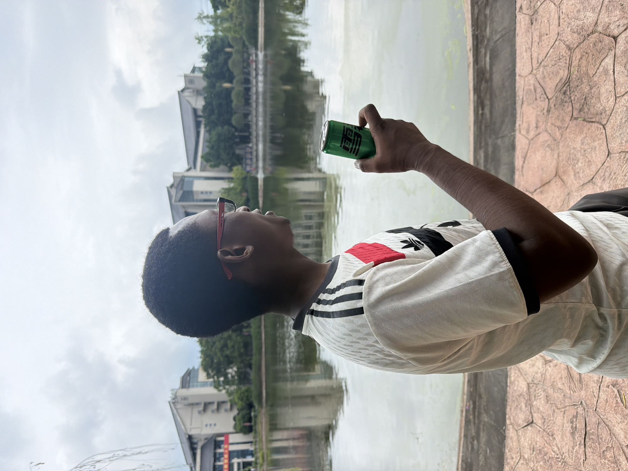 An individual, identified as a young Black male wearing glasses and a white, black, and red soccer-style jersey, stands outdoors holding a green can of soda in his right hand. He is positioned with his back to the viewer, looking to the right towards a body of water. Behind the water, a city skyline is visible, featuring several buildings and lush green trees. The sky is overcast with clouds. The ground in the foreground is paved with reddish-brown interlocking stones. This scene is located in Jiangjin, China.