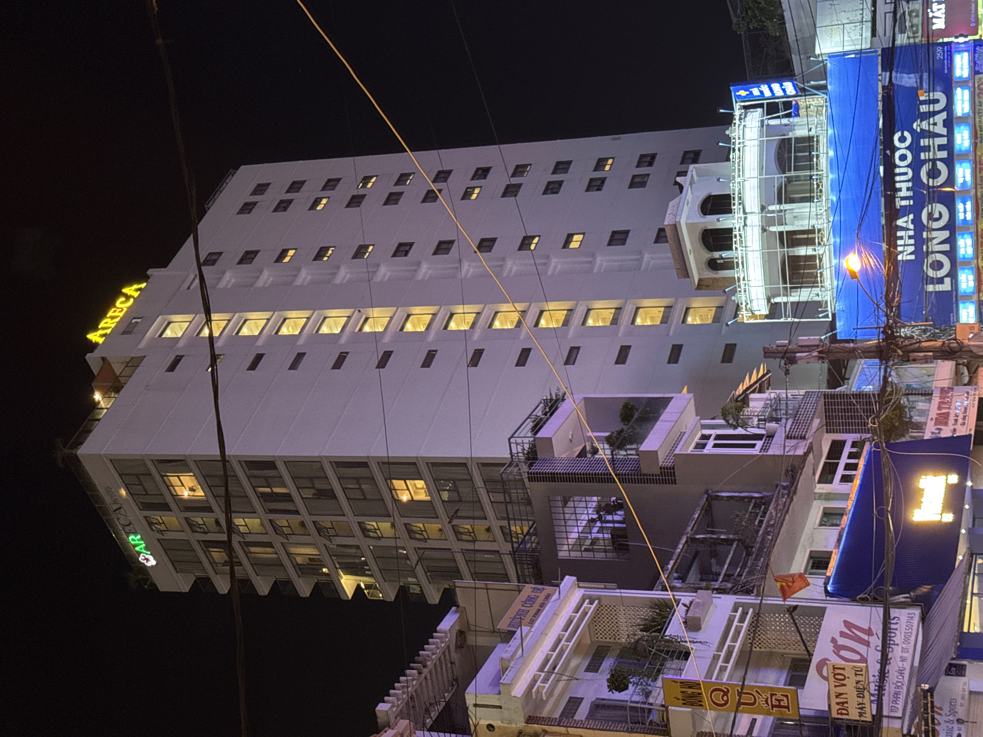 The image shows a nighttime street view in Nha Trang, Vietnam. A tall, white building with numerous illuminated windows dominates the scene. The building has a green and yellow neon sign at the top that reads "ARECA". Below the tall building, there are other structures with varying levels of illumination and signage. One prominent sign in blue reads "NHA THUOC LONG CHAU", indicating a pharmacy. There is a Vietnamese flag visible on a balcony. Overhead, there are power lines crisscrossing the frame. The overall impression is of a busy urban environment at night.