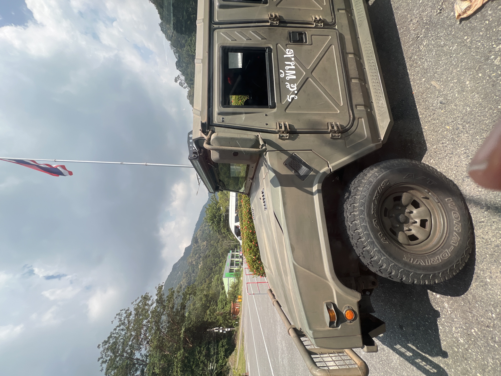 A military-style vehicle, painted olive drab, is parked on an asphalt road. The vehicle has a brush guard on the front and is partially obscured by a hedge of red flowers. The side of the vehicle has white Thai script. In the background, there is a paved road, a street light, a modern green and white building, and a white van. Further in the background, lush green mountains rise under a cloudy sky. A Thai flag is flying on a flagpole to the left of the vehicle. The scene appears to be in a rural or semi-rural area.