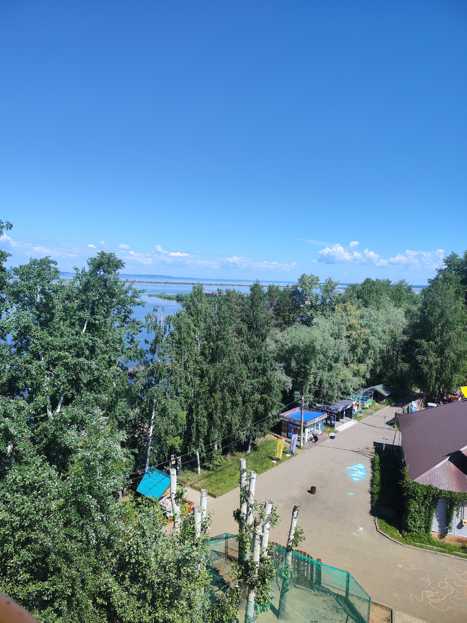 The image depicts an outdoor scene with a clear blue sky above a body of water and a tree-lined landscape. In the foreground, lush green trees, some with white bark, dominate the view. Beyond the trees, a wide expanse of blue water stretches out, with a thin line of land visible in the distance. A few scattered white clouds dot the sky.
Closer to the viewer, nestled amongst the trees and to the right of the water, is a small, developed area with a few structures. A brown-roofed building with a light-colored wall covered in ivy is visible. Adjacent to it is a paved area with some chalk drawings on the ground, including a white wavy line and some blue chalk marks that resemble clouds. A single dark cylindrical object, possibly a trash can or a small post, stands in the middle of the pavement.
Further into the scene, several small colorful kiosks or stalls are visible, some with blue and red awnings. These appear to be part of a recreational area or marketplace. A paved path or road winds through this section, connecting the various structures. In the background, more trees and vegetation suggest a natural or semi-natural environment surrounding the developed area. The overall impression is that of a sunny day in a location that combines natural beauty with some recreational facilities.