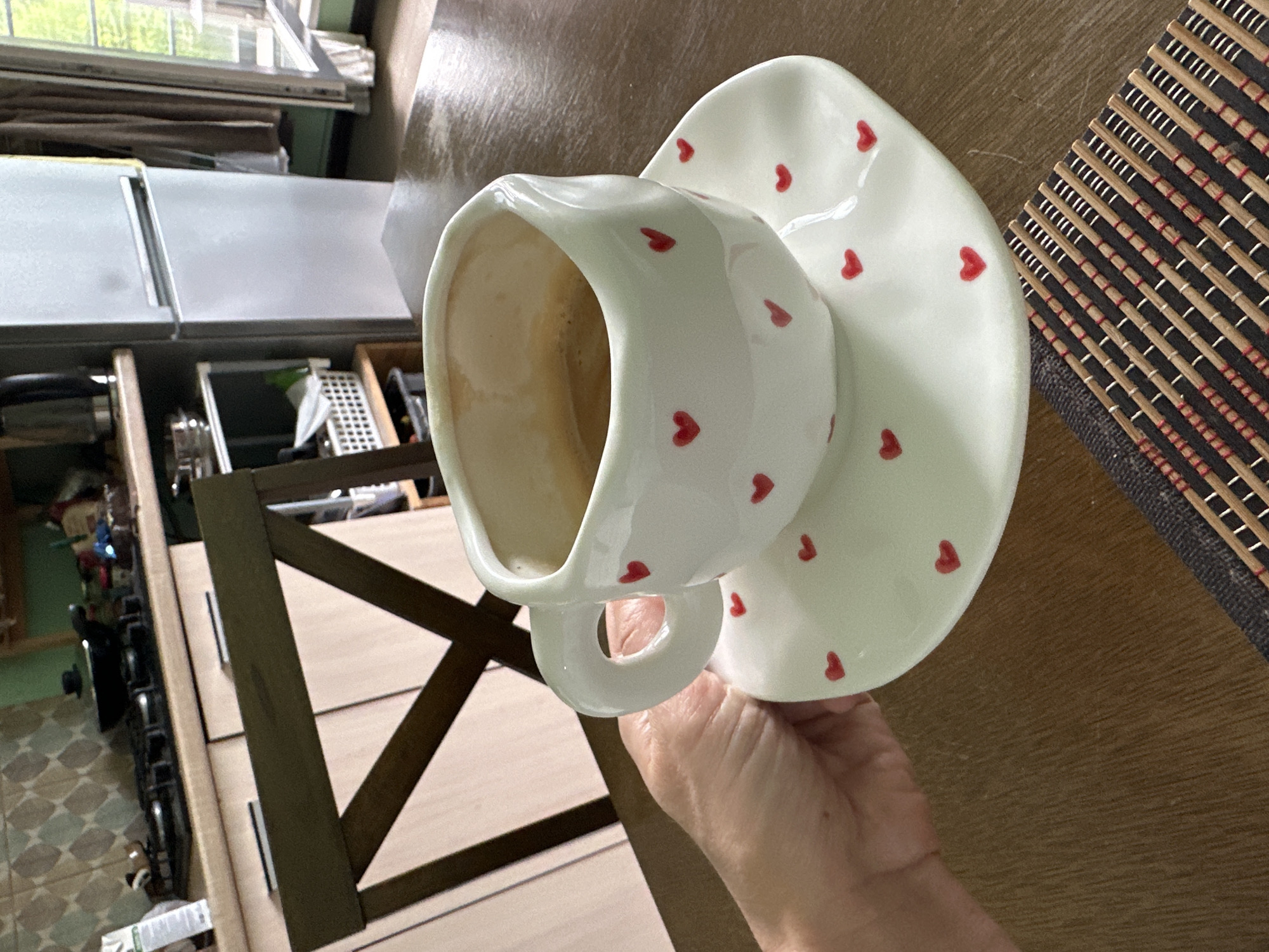 An over-the-shoulder shot shows a person holding a white coffee cup with red hearts. The cup is on a matching saucer, also adorned with red hearts. The cup is partially filled with coffee. The subjects are on a wooden table, with a placemat in the foreground. In the background, a kitchen scene is visible, including cabinets, a stovetop with a pan, a refrigerator, and a window. The lighting suggests it is daytime.