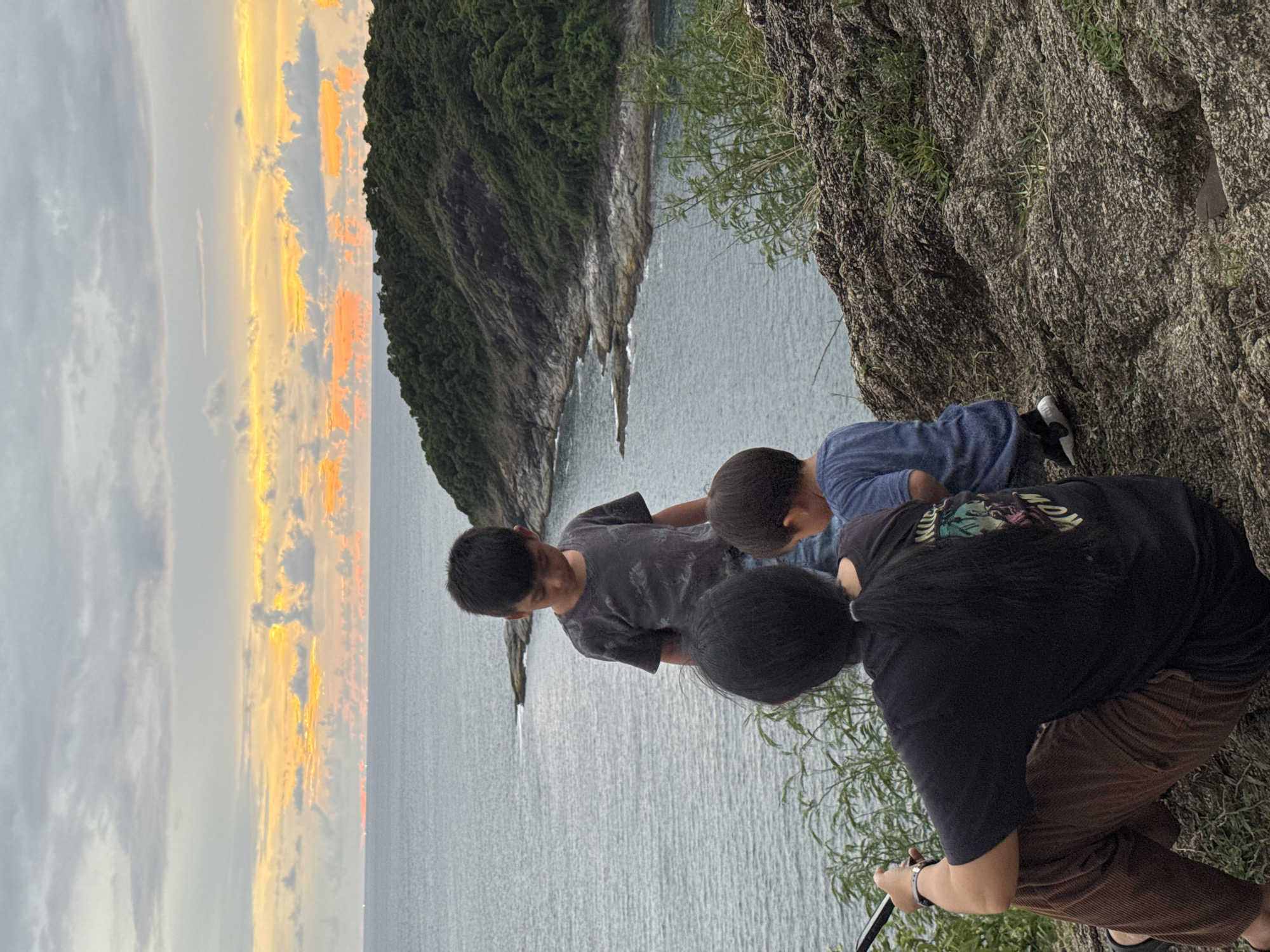 This image depicts a family enjoying a sunset view at Promthep Cape in Rawai, Thailand. In the foreground, a person with long black hair tied in a ponytail is seated on a rocky outcrop, facing away from the camera and holding a mobile phone. To their right and slightly behind, two young boys are standing. The older boy, wearing a dark t-shirt, looks down, while the younger boy, in a blue shirt, is turned away. The middle ground features the calm, grey sea meeting a lush green, tree-covered peninsula. The horizon is dominated by a dramatic sky with streaks of orange and yellow clouds, illuminated by the setting sun. The overall scene is one of peaceful observation of the natural landscape.