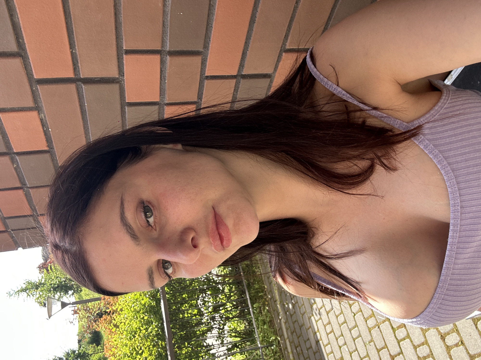 A young woman with long, dark brown hair and fair skin is pictured in a close-up selfie. She is wearing a light purple ribbed tank top that reveals her chest. Her eyes are looking directly at the camera, and her lips are slightly parted. Her skin appears to have some redness and blemishes. The background shows a brick wall with varying shades of brown and orange, and to the left, there is greenery with some trees and a lamp post. The ground is paved with light-colored bricks. The lighting suggests it is daytime, with sunlight casting shadows.