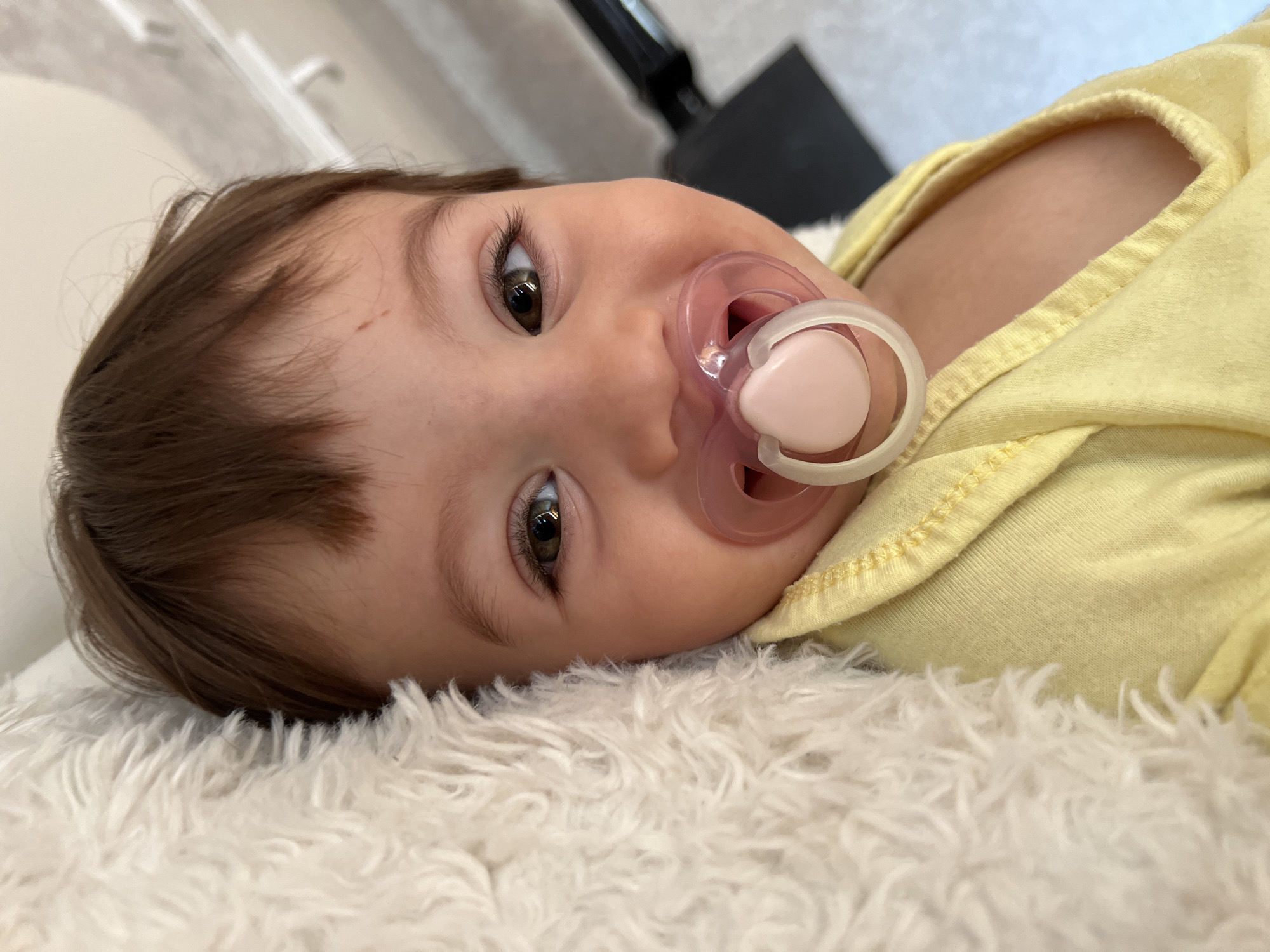 A close-up, eye-level shot captures a baby's face, positioned to the right of a white fluffy blanket that occupies the left side of the frame. The baby, facing forward, has dark hair and brown eyes, with a pink pacifier in its mouth. It wears a yellow top. The background consists of a bright, out-of-focus surface.