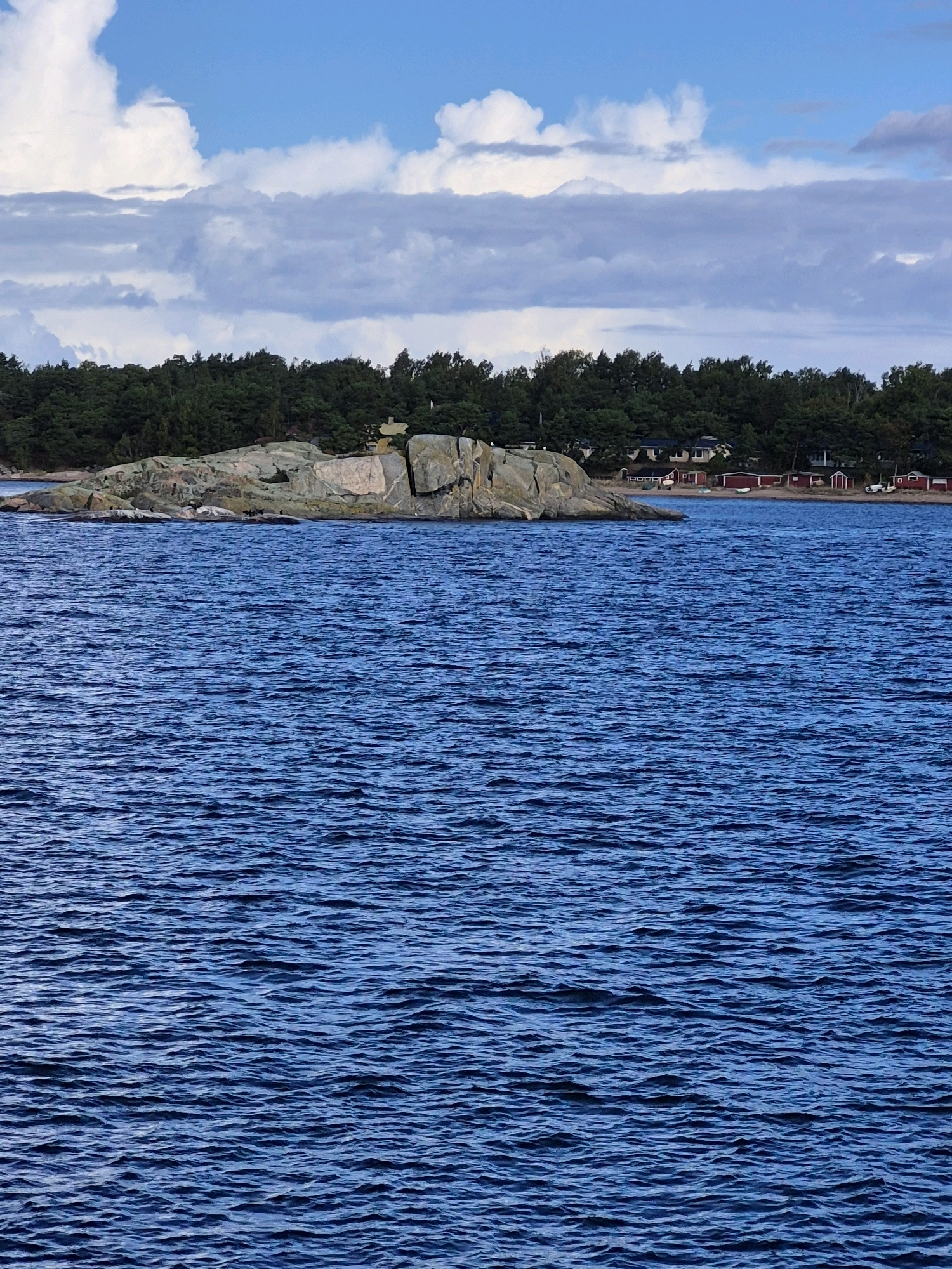 A wide shot captures a body of deep blue water, its surface rippled by gentle waves, in the foreground. In the mid-ground, a rocky island juts out from the water, topped with a scattering of green trees. Beyond the island, a tree-lined shore is visible, with a few red buildings and houses nestled amongst the trees. The sky above is a mix of bright blue and dramatic, billowy white and grey clouds. The scene is reminiscent of the coastal areas of Hanko, Finland, characterized by its archipelago and rocky islands.