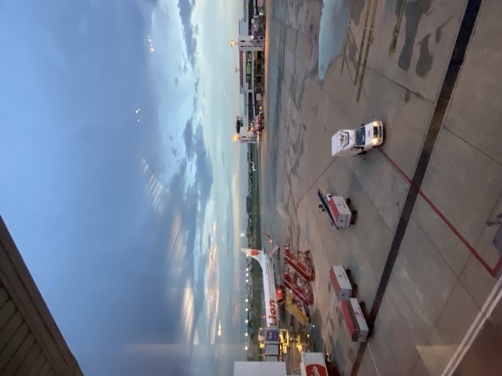 The image depicts an airport scene at dusk. A Lion Air aircraft is parked at the gate, with stairs and ground service equipment nearby. Other vehicles are visible on the tarmac. The sky is cloudy with visible light. The location is likely Suvarnabhumi Airport in Bangkok, Thailand, given the presence of a Lion Air aircraft, a budget airline based in Southeast Asia. There are no people visible in the image. The image is taken from inside the airport terminal looking outwards.