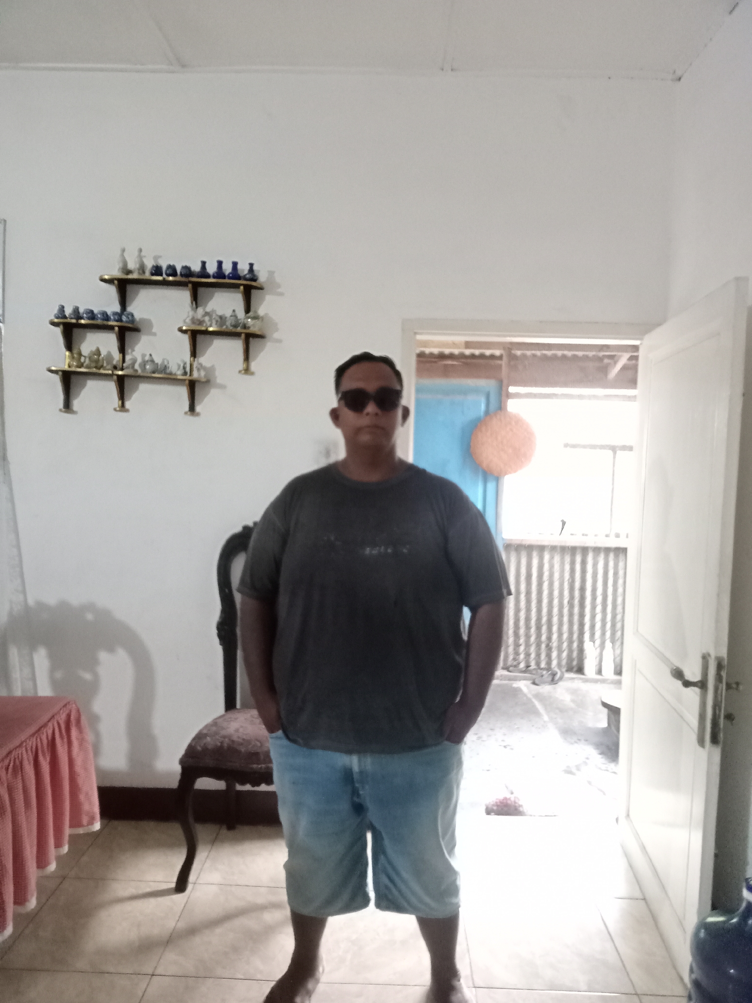 A full-body, medium shot captures a person standing in an indoor setting with an open doorway leading outside. The individual, who appears to be male, is wearing dark sunglasses, a dark grey t-shirt, and light blue denim shorts. His hands are tucked into his pockets, and he is facing the camera. To the left of the person, a chair with an ornate carved back and upholstered seat is visible. Further to the left, a table covered with a pink tablecloth that has a ruffled hem is partially in frame. The walls are painted white, and a series of small decorative bottles and objects are displayed on tiered shelves mounted on the wall behind the person. The doorway reveals a glimpse of an outdoor area with corrugated metal siding and some indistinct structures. The flooring inside appears to be tiled. The image is taken from a slightly low angle. The location is identified as Muliorejo, Indonesia.
