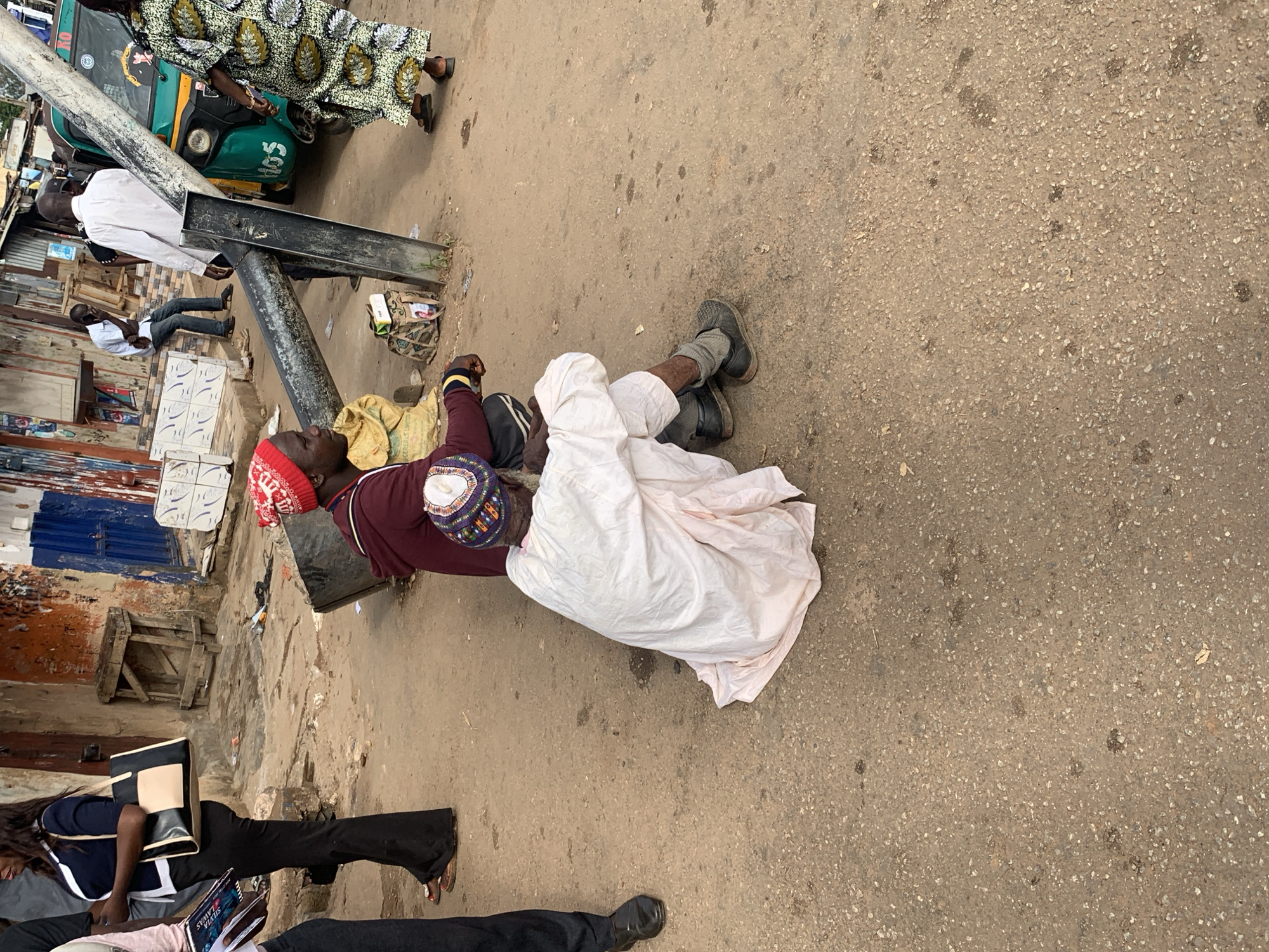 In Gura Top, Nigeria, a busy street scene unfolds. A woman in a navy blue and white top and black flared pants, carrying a black and cream bag, stands on the left. Next to her, a person in a maroon hoodie and a red beanie sits on a yellow sack. To their right, another individual, dressed in a white robe and a colorful cap, is seated on the ground. A metal pole, likely part of a street light or signpost, runs diagonally across the frame. In the background, a green and yellow auto-rickshaw is parked. The ground is a dusty, textured surface. People are present, some appear to be vendors or pedestrians in a marketplace setting.