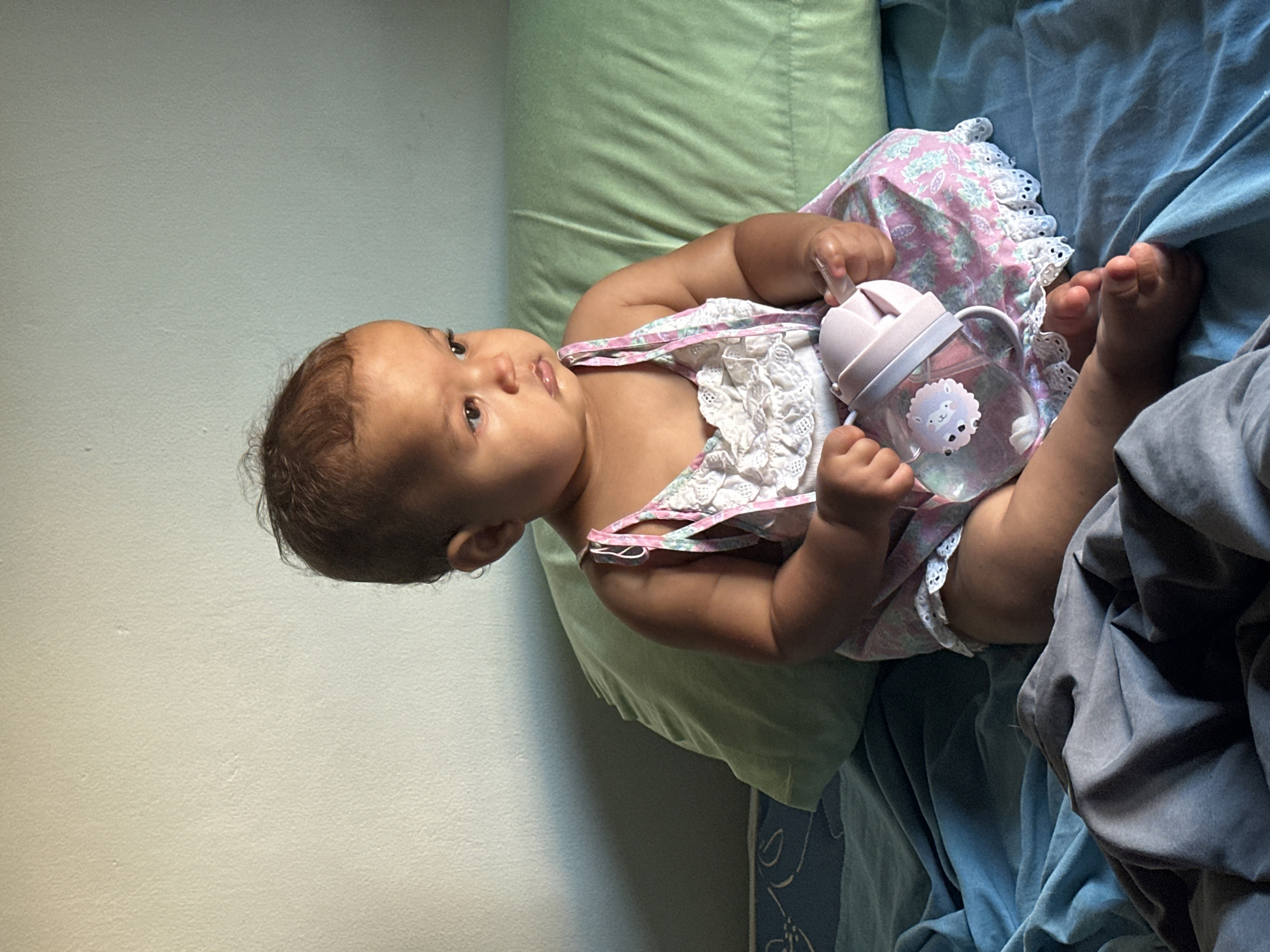 A baby lies on a bed with a green pillow and blue sheets. The baby is wearing a dress with pink and white lace. The baby is holding a pink and clear bottle with a sheep sticker. The background is a light wall. There is no evidence of North Kuta, Indonesia.