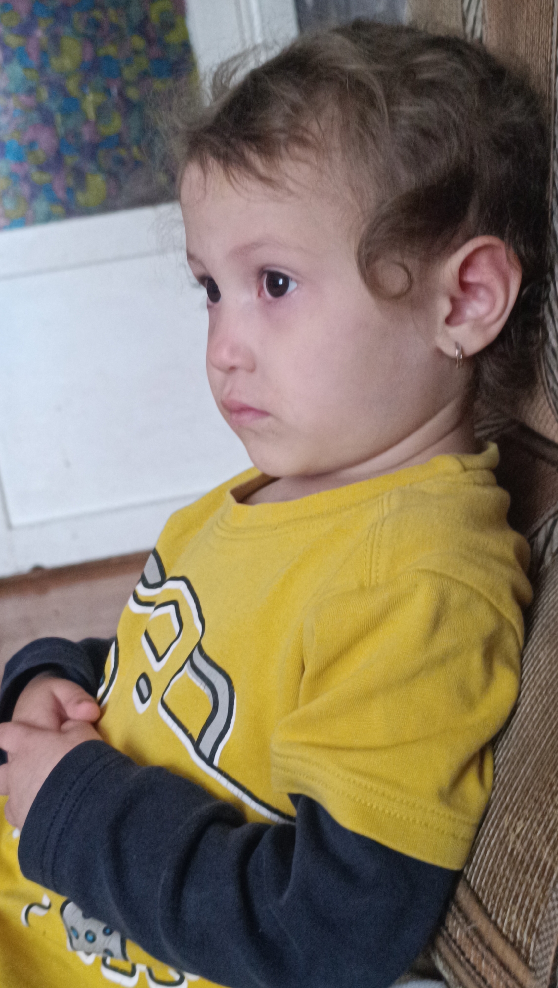 A medium shot captures a young child with curly brown hair, an earring in their left ear, and dark eyes, looking to the left of the frame. The child is wearing a yellow long-sleeved shirt with a dark blue undershirt. The child's arms are crossed. The background shows a patterned wall on the left and a textured surface on the right, possibly a couch or chair. The setting appears to be indoors. There is no discernible information in the image to indicate a specific location in Karsha, Russia.