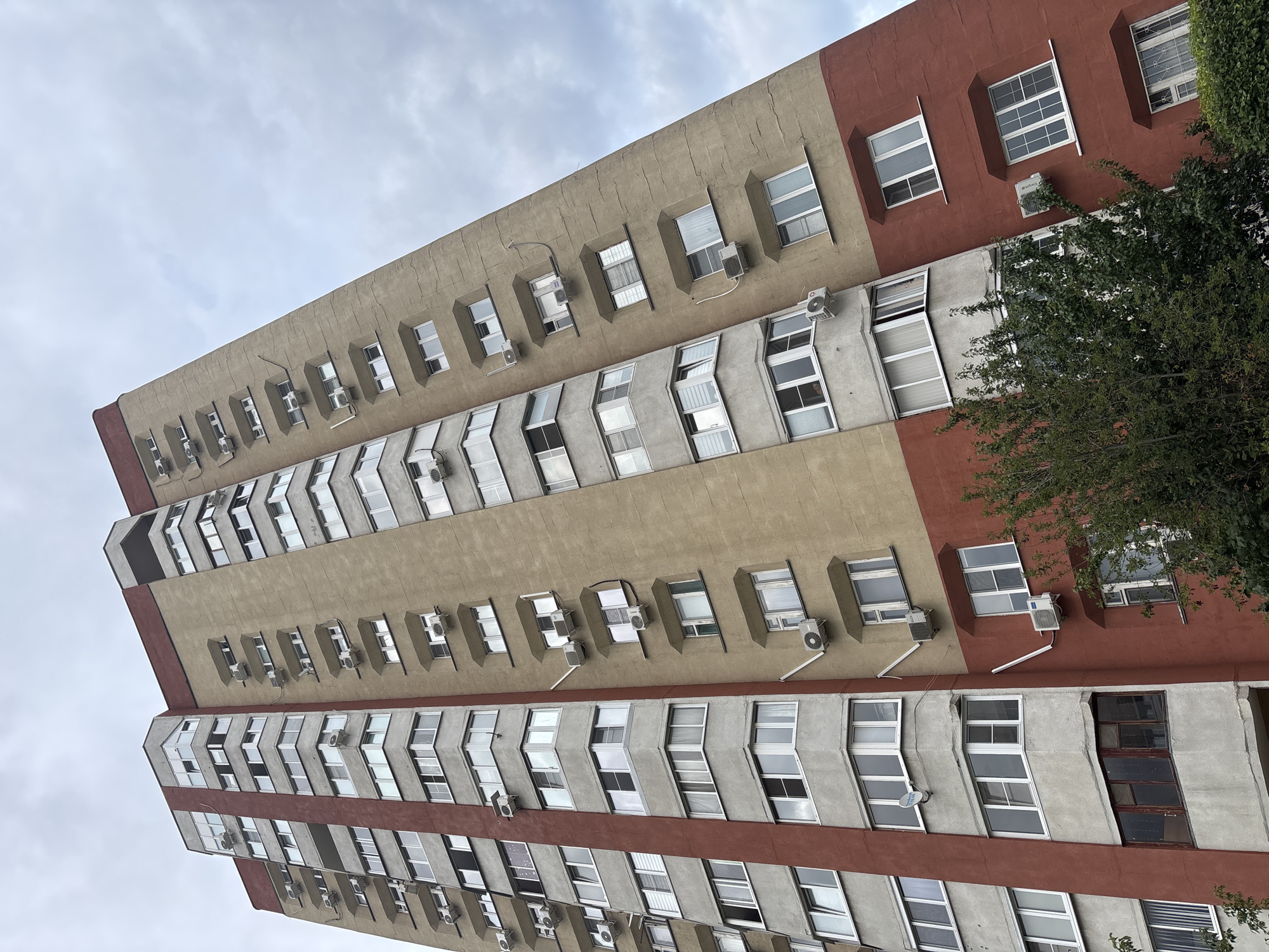 The image shows a multi-story apartment building from a low-angle perspective against an overcast sky. The building has a distinct color scheme, with the lower section painted a dark red and the upper sections a beige or light brown. Many windows are visible on each floor, some with air conditioning units mounted outside. The building appears to be in an urban environment, with trees and foliage visible at the base of the structure. Based on the architectural style and the presence of specific details, this building is likely located in Tolyatti, Russia.