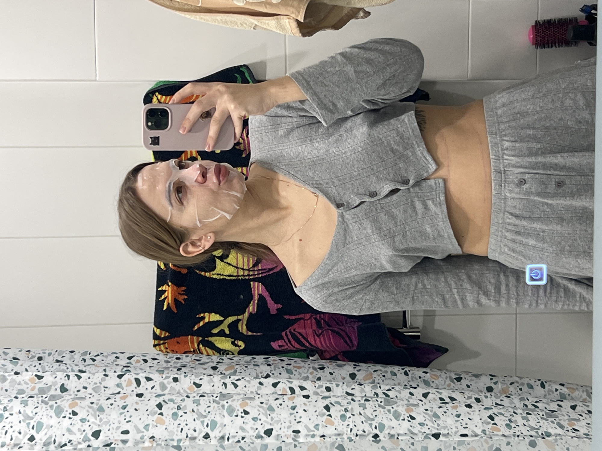 A young adult female is depicted in a mirror selfie taken within a bathroom located in Krasnoyarsk, Russia. She has light brown hair, secured back, and is wearing a white sheet face mask on her face. Her attire consists of a grey ribbed two-piece loungewear set: a cropped, long-sleeved, button-front top, and matching bottoms, exposing her midriff. She wears a gold-colored necklace and has visible piercings in her right ear. A tattoo is partially visible on her left torso. The female holds a light pink smartphone, featuring a black cat sticker, to capture her reflection; her head is slightly turned to the right with pursed lips. The bathroom features white tiled walls. A white shower curtain with a multi-colored terrazzo pattern (grey, teal, peach) is present on the left. Behind her, a dark towel with orange, yellow, and magenta patterns hangs on a rail. A lighter, beige-colored towel is visible on the right. A pink and black hairbrush rests on a counter surface in the lower right, and a glowing blue power icon is visible on the mirror's surface, indicating a functional element.