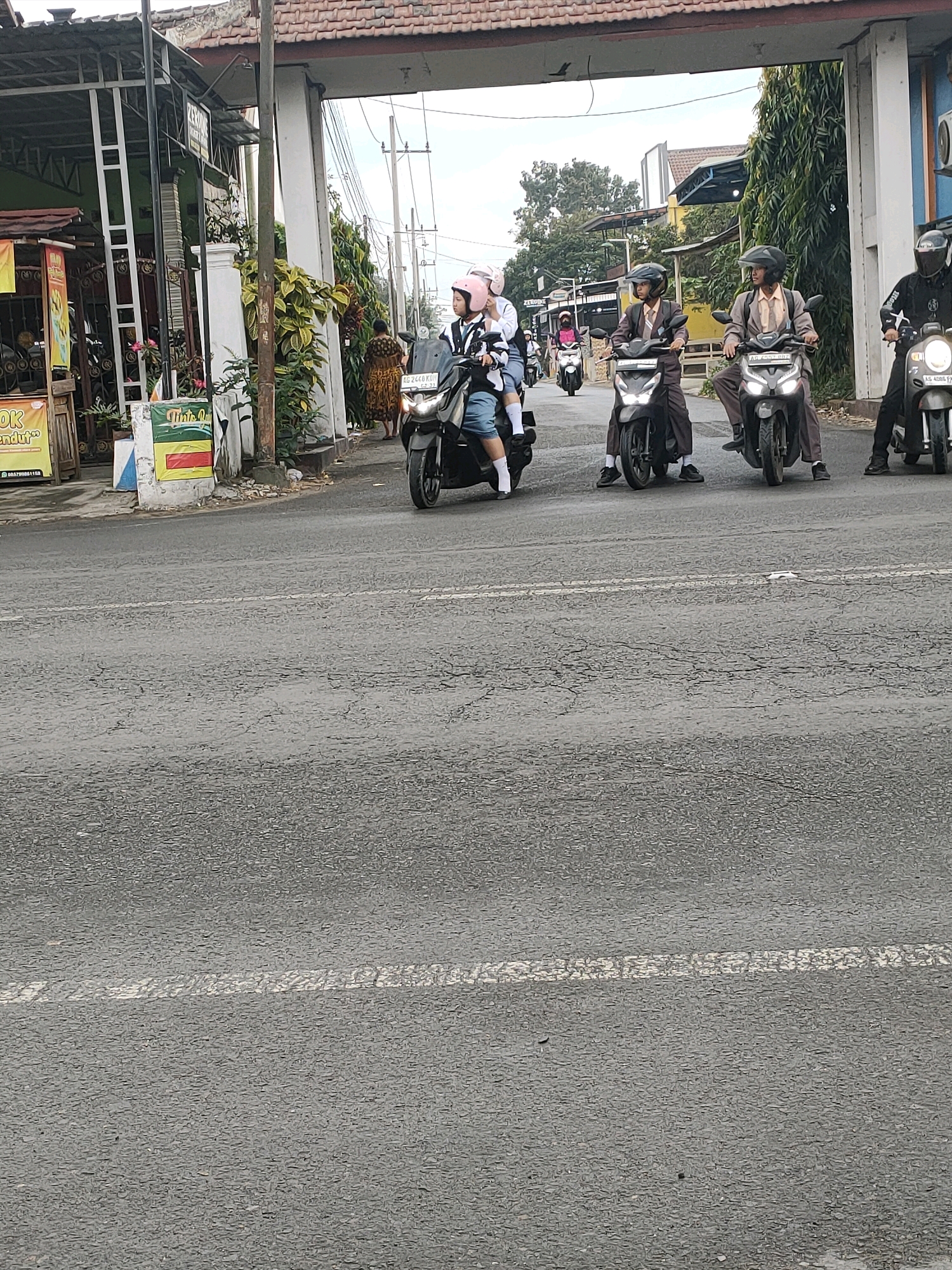 In the city of Kanigoro, Indonesia, several individuals are riding motorcycles under an arched gateway. The scene takes place on a paved road with white lane markings. On the left side of the road, a stall with the text "OK KENDURI" is visible. Several people are on motorcycles in the foreground and middle ground. Two individuals in brown school uniforms and helmets are riding parallel to each other. To their right, another person wearing a black helmet and jacket rides a motorcycle. To the left, a person in a pink helmet is riding a motorcycle, with another person behind them, also wearing a helmet. In the background, more motorcycles and pedestrians can be seen. The environment appears to be a street with buildings and vegetation lining the sides, under an overcast sky.