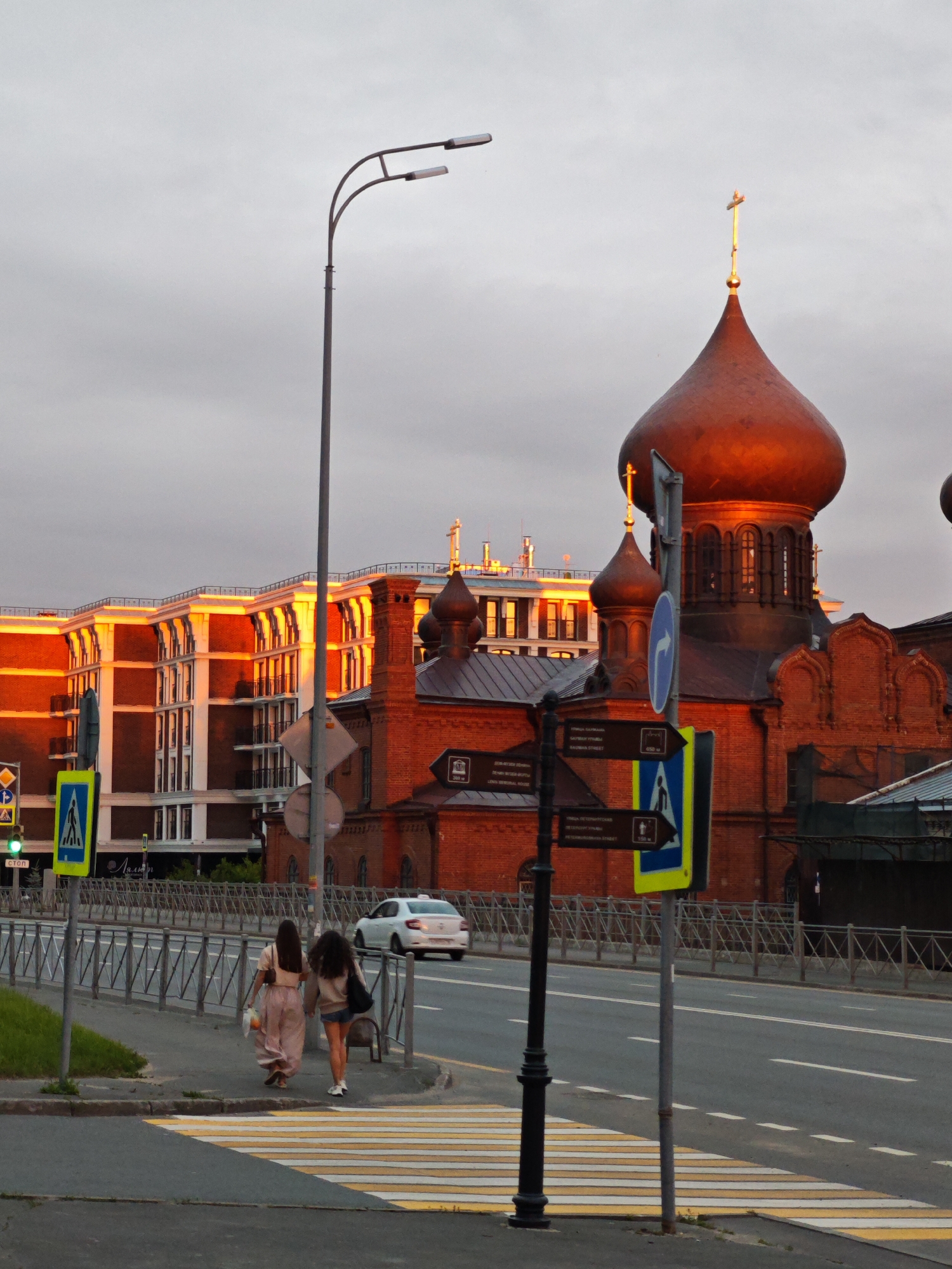 The image depicts a street scene in Kazan, Russia, during what appears to be sunset, casting a warm glow on the buildings. In the foreground, two young women walk away from the camera on a sidewalk, crossing a pedestrian zebra crossing. One woman wears a light pink dress and carries a bag, while the other has on a light-colored top and denim shorts, also carrying a bag.
To the right of the women, a street lamp stands tall, and further down the street, a white car drives away from the viewer. Several street signs are visible, including traffic signs indicating pedestrian crossings and directional signs with arrows.
Dominating the background is a traditional Russian Orthodox church with a prominent copper-colored dome topped with a golden cross. Adjacent to the church is a modern building with a red brick facade and white architectural elements, reflecting the sunset's light. The sky is overcast with a diffused light. A metal fence separates the sidewalk from the road.