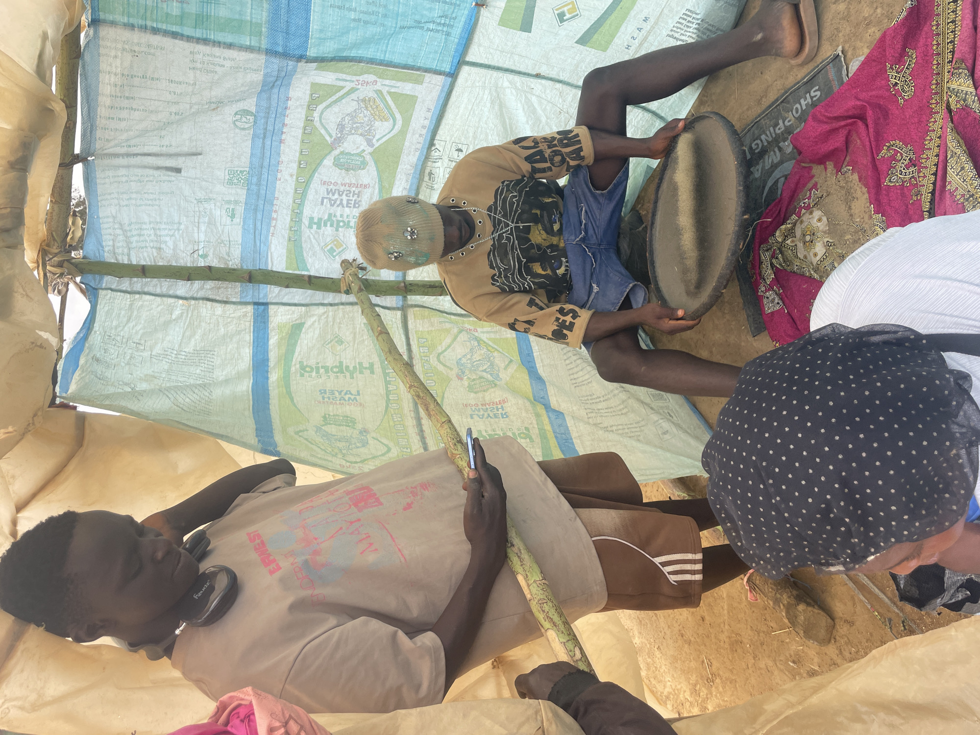 Captured indoors, the image presents a group of individuals within a makeshift shelter constructed from repurposed fertilizer bags, bearing the brand "Hybrid."
Centered, a seated person, adorned with a decorated beanie and a patterned top, holds a circular object, possibly a drum or tray. To the right, partial views of another person with a polka-dotted head covering and a piece of pink fabric are seen.
On the left, a man in a graphic t-shirt and headphones holds a slender, green cane or stick, also interacting with a phone.
The shelter's interior reveals a dirt ground and minimal furnishings, adding to the sense of poverty. A "SHOPPING AMA" sign lies near the seated person. The image appears to be a candid snapshot of everyday life in Maiyanga, Nigeria.