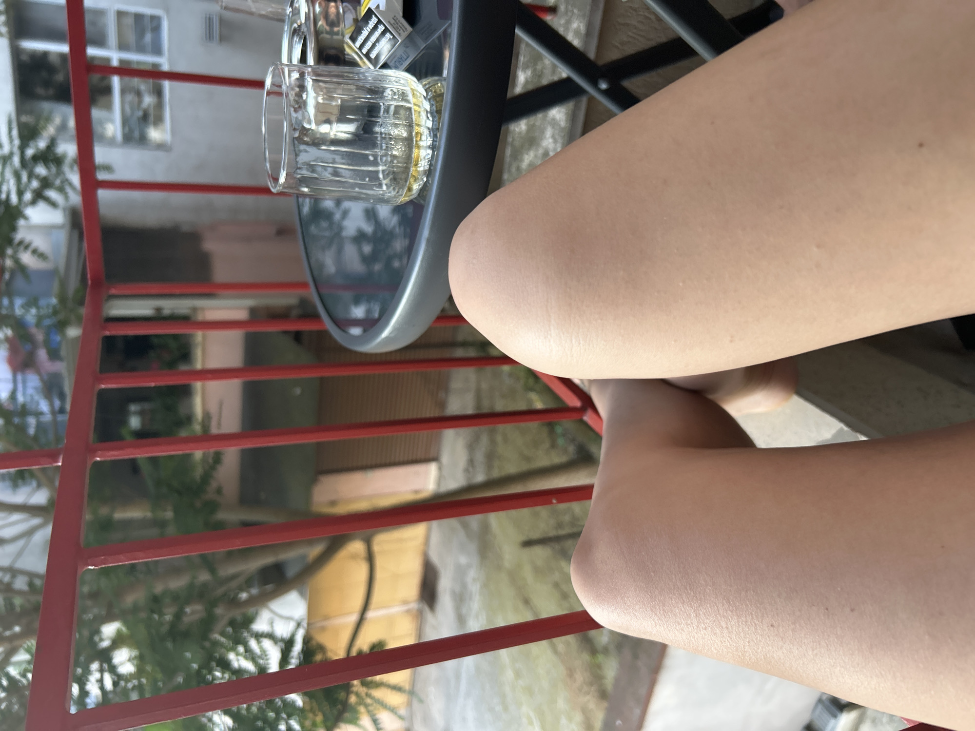 The image shows a close-up of a person's bare legs, specifically their thighs and knees, positioned in the foreground. They are sitting on a balcony or patio with a red railing visible. A round, dark-colored table with a glass of amber liquid and a pack of cigarettes rests on its surface. In the background, trees and buildings are visible, suggesting an urban setting. The light suggests it is daytime. The specific location of Burgas, Bulgaria, is not discernible from the visual information in the image itself.