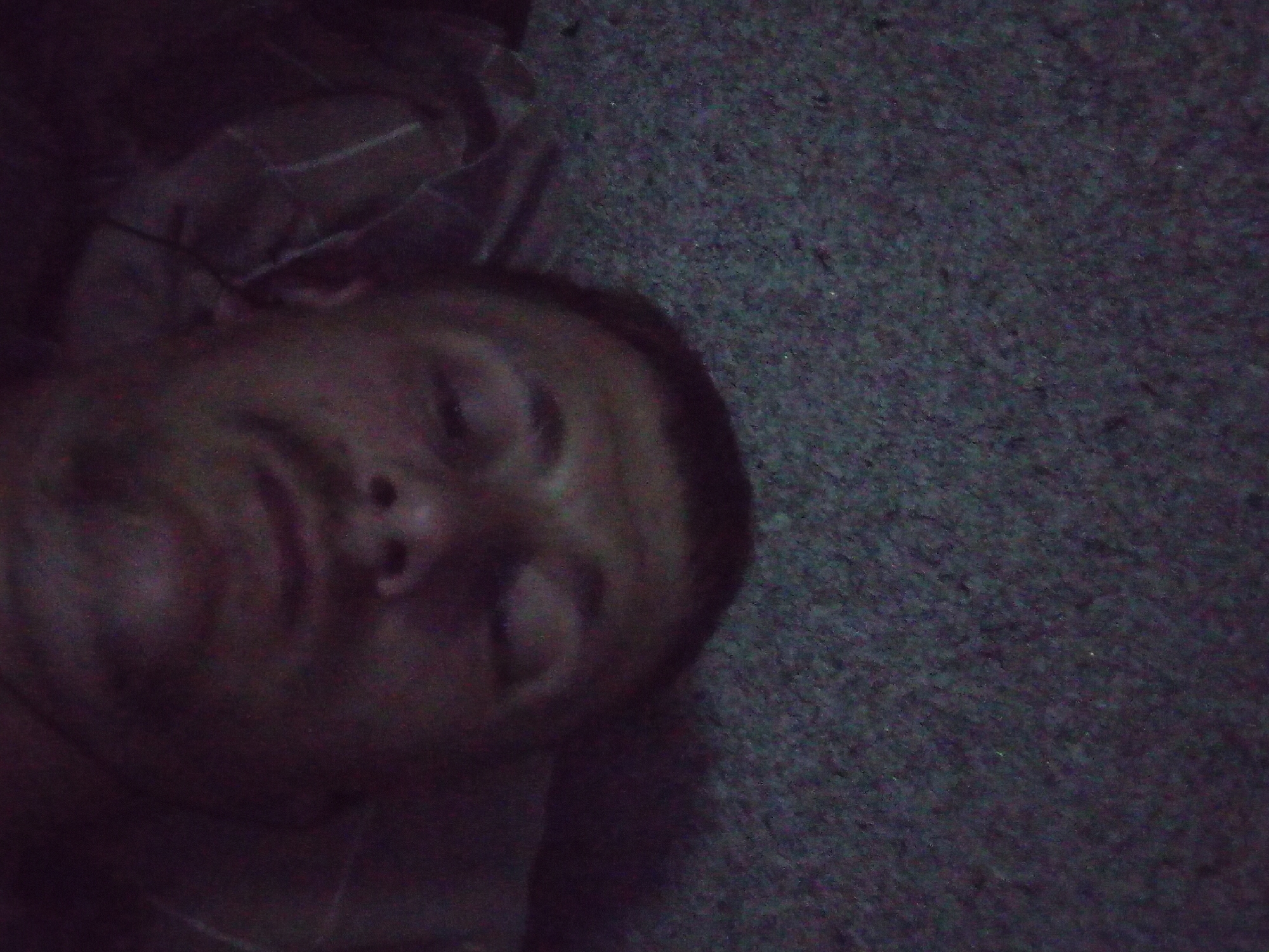 A close-up, low-light, tilted shot features a person lying down on a textured, dark gray surface, possibly carpet or a similar fabric. The person's face is partially visible, with their eyes closed, suggesting they are sleeping or resting. The lighting is dim, casting deep shadows and obscuring some details of the face and surroundings. To the left, a patterned fabric, likely clothing or bedding, is visible, with lighter stripes on a darker background. The overall mood is subdued and intimate due to the low light and the subject's relaxed state. No text or specific location cues are discernible in the image.
