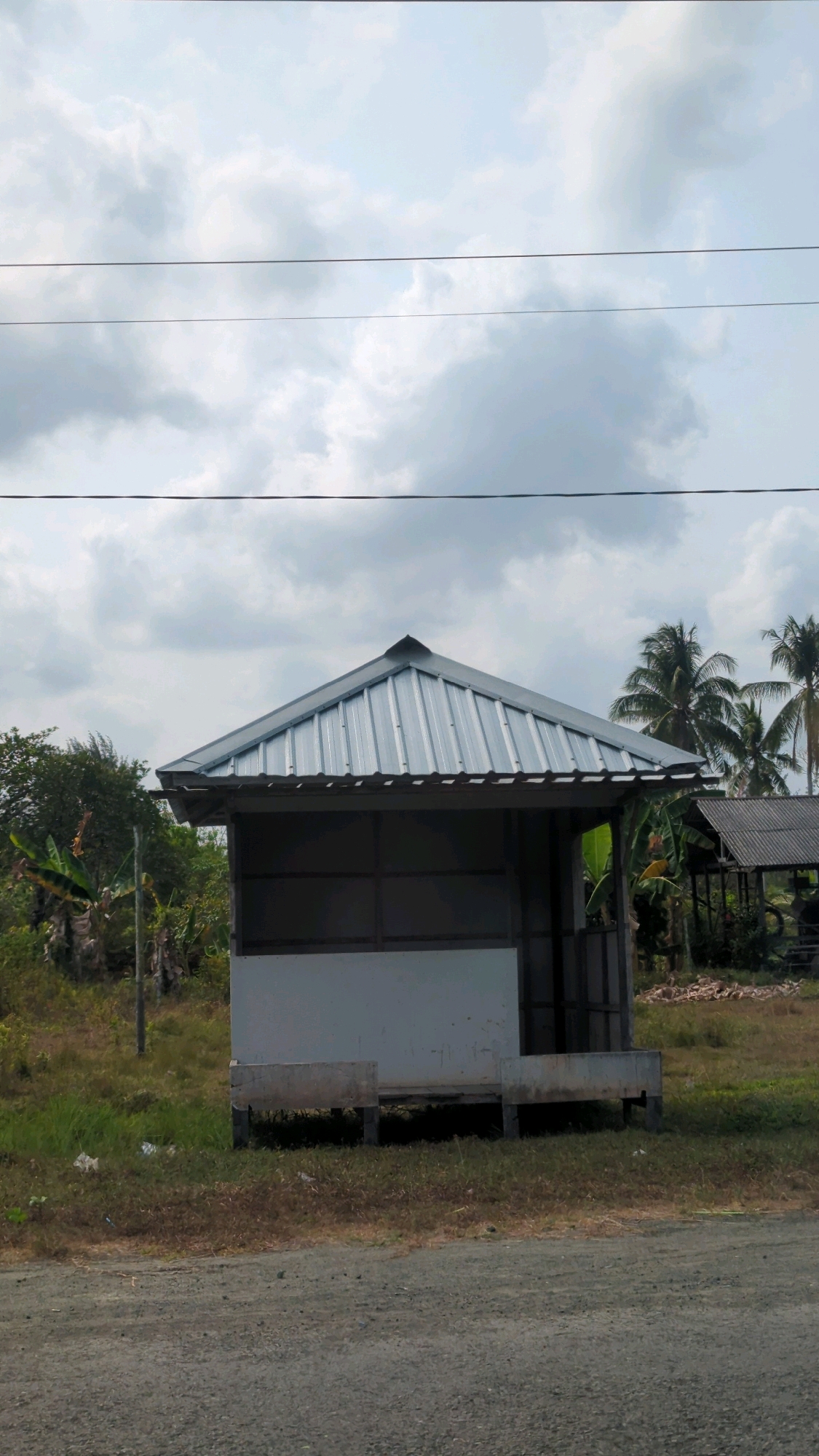 This is a photograph of a small, open-sided structure with a corrugated metal roof, situated in a rural area. The structure appears to be elevated on wooden stilts. In front of the structure, there is a low concrete planter box. The area surrounding the structure is grassy and somewhat overgrown, with some scattered debris. Behind the structure, there are palm trees and other vegetation, including what appears to be banana trees. Further in the background, another similar structure with a corrugated roof is visible. The sky is cloudy. The photograph seems to have been taken from the side of a dirt or gravel road. The location is identified as Muara Pagatan, Indonesia. There are no people visible in the image.