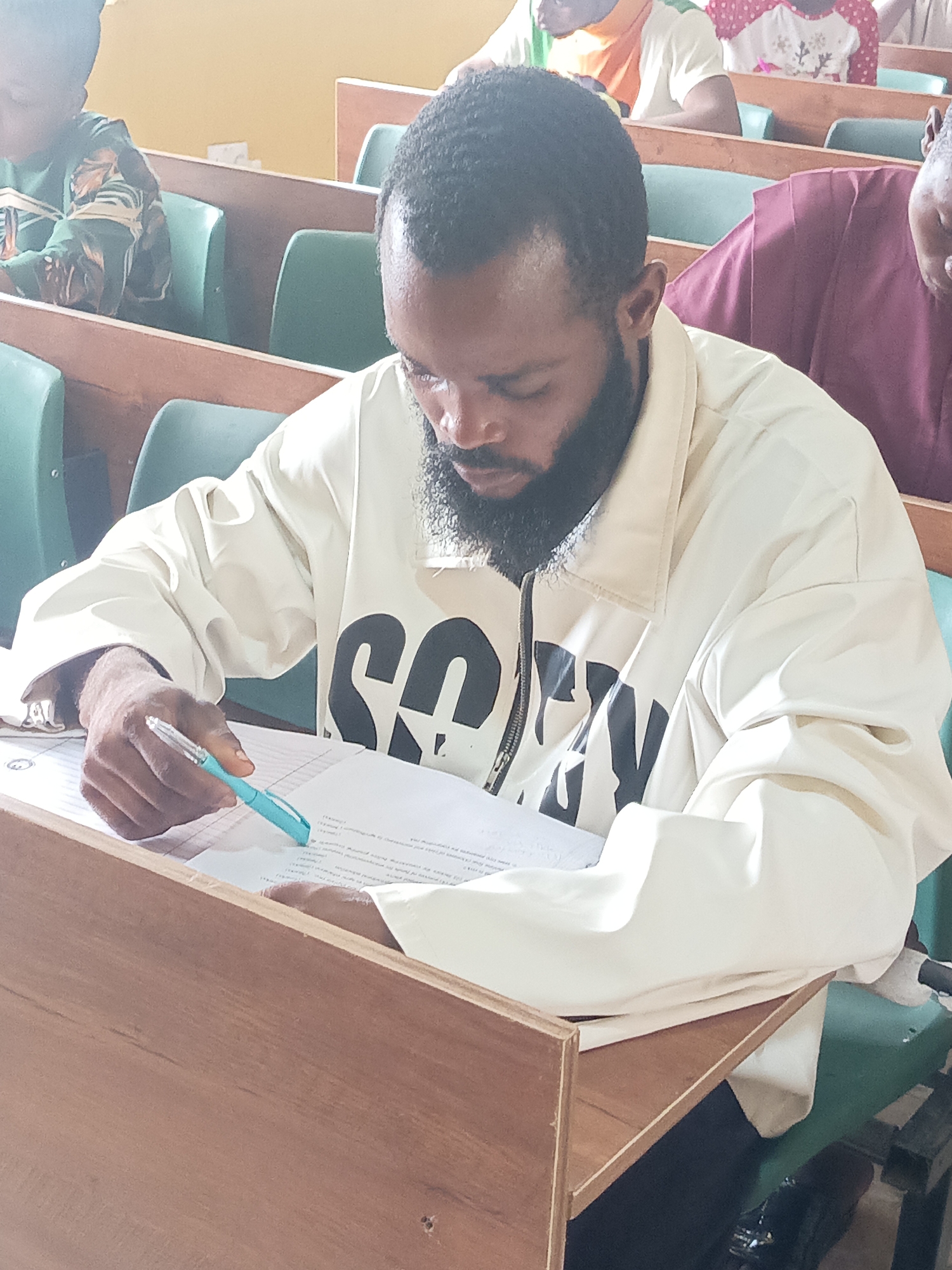 This image captures an indoor scene, likely a lecture hall, classroom, or examination venue in Akwanga, Nigeria. The foreground is dominated by a man with dark skin, short dark hair, and a full dark beard. He is wearing a light-colored (possibly beige or off-white) jacket with large, dark, block letters on the front, which appear to spell "SOF" and a partial fourth letter. He is seated at a wooden desk with a green chair, intently focused on papers laid out before him. His right hand holds a light blue pen, poised over the documents, suggesting he is writing, taking an exam, or reviewing material.
In the background, several other individuals are partially visible, also seated in similar wooden desks with green chairs arranged in rows. To the man's left, a person wearing a green patterned top is visible. To his right, another person is seen in a maroon or purple garment. Further back, another individual wears a white top with red polka dots. The setting, with its multiple rows of uniform desks and seating, points to a formal environment for learning or assessment. The man's posture and gaze convey deep concentration. The papers on his desk contain small printed text, with some structured elements like numbers in parentheses, typical of questions or prompts. The bright lighting suggests it is daytime.