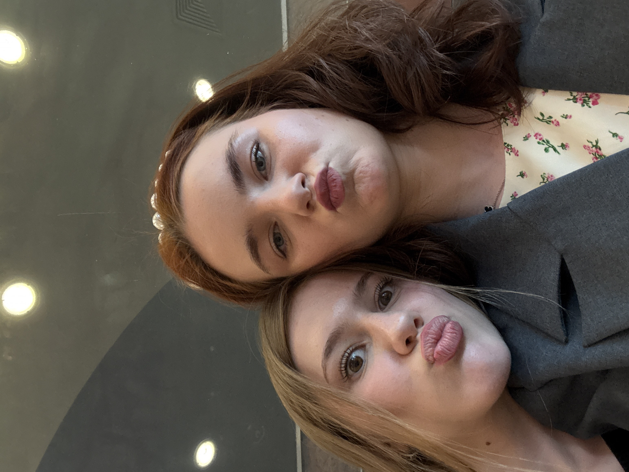 Two young women pose closely for a selfie in an indoor setting. The woman on the left has reddish-brown hair, styled with a pearl-like headband. She wears a light-colored top with a delicate pink floral pattern beneath a dark grey or black blazer. Her eyes are light and open, looking directly at the camera, while her lips are pursed in a "duck face" expression. A small black heart-shaped pendant is visible on her necklace.
Leaning against her, on the right side of the frame, is a woman with long blonde hair, also wearing a dark grey or black blazer. Her brown eyes are wide open and directed towards the camera, and her lips are similarly pursed in a "duck face." Both women exhibit fair complexions and soft makeup, with prominent eyelashes and eyebrows.
Their heads are touching, indicating a close and friendly interaction. The background is dark and features several bright, circular reflections, suggesting artificial lighting on a glossy or reflective surface, possibly a window or a polished wall. Faint geometric patterns are visible in the upper left background. The overall mood conveyed is playful and casual. There is no visible text in the image. The setting is Saint Petersburg, Russia.