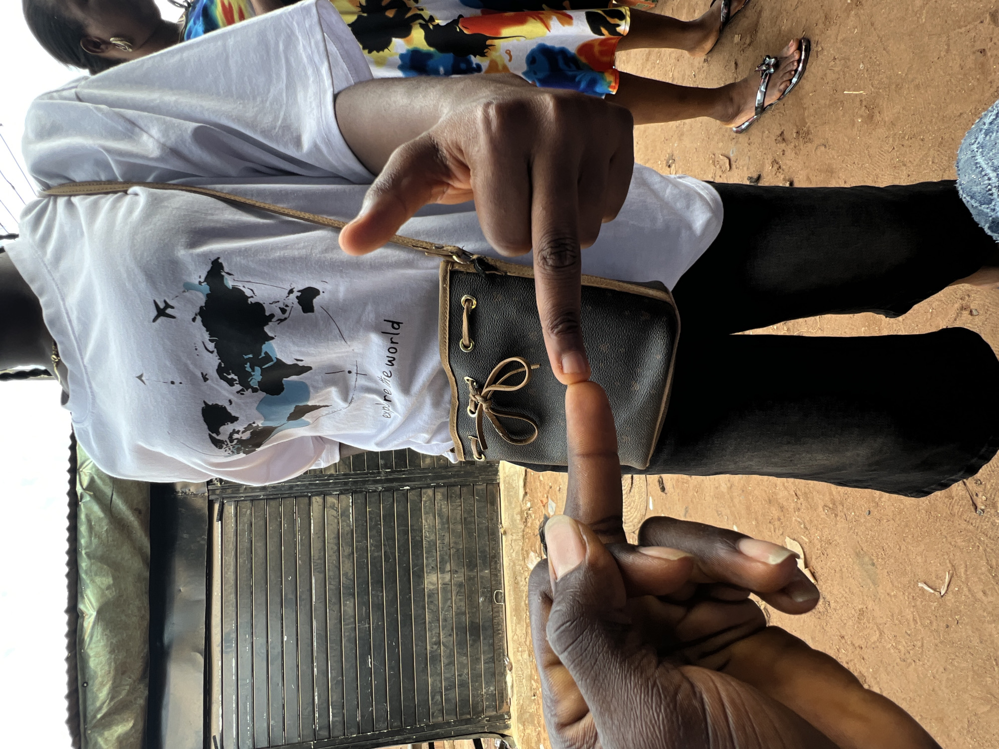 In Nkpor, Nigeria, two individuals are positioned outdoors. The subject in the foreground has dark skin and is wearing a white t-shirt with a world map graphic and the words "explore the world." They are holding a small, dark brown purse with a drawstring closure. Their right hand is raised, showing a thumb and index finger pointing gesture, while their left hand is visible in the lower left corner, with fingers extended. A second individual, partially visible to the right, wears a colorful patterned dress and flip-flops. The background consists of a dark, corrugated metal structure, possibly a shop or stall, with a green tarp overhead. The ground is covered in reddish-brown dirt.