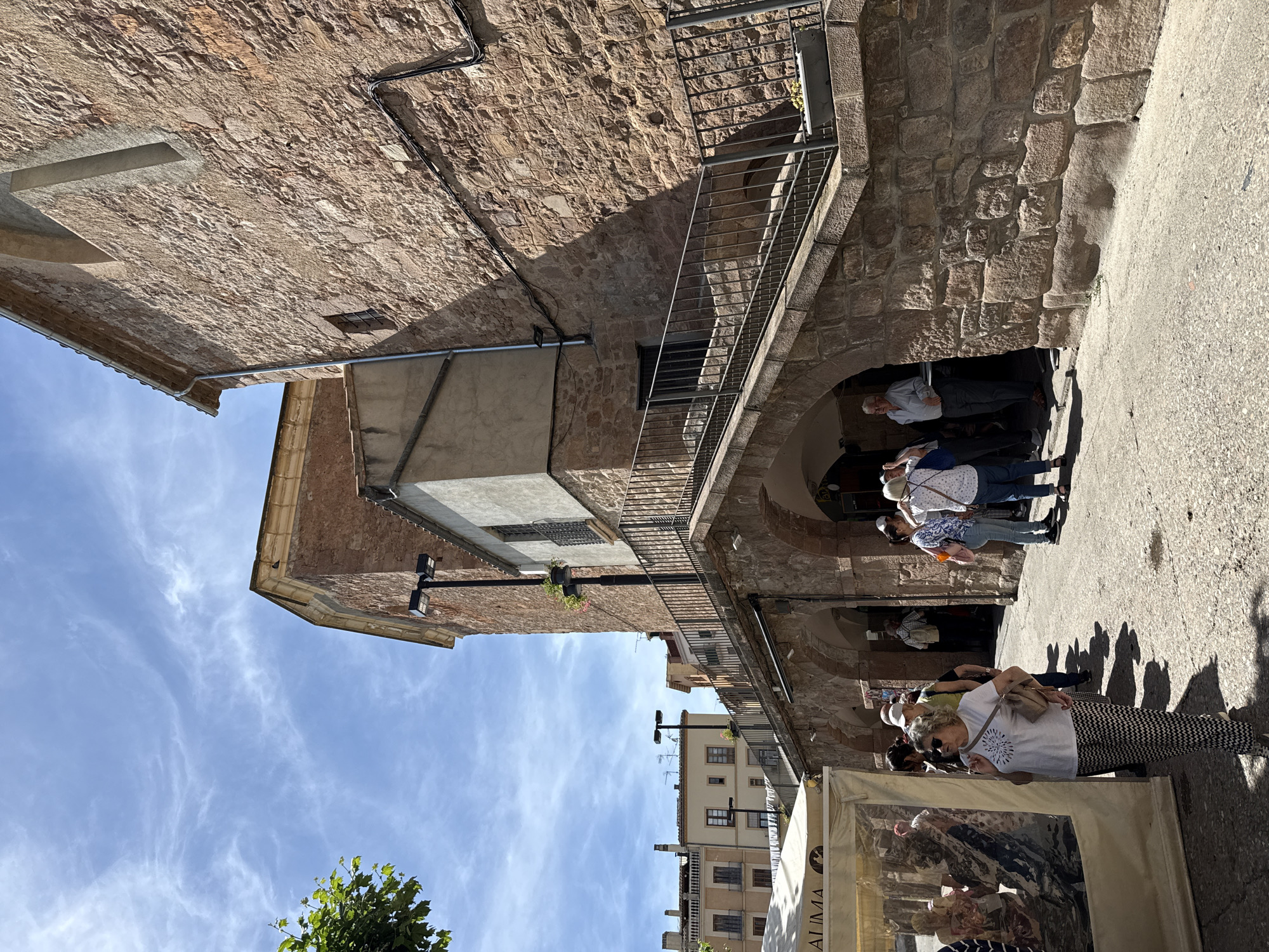 The image depicts a street scene in the historic town of Cardona, Spain, likely near the medieval castle or old town. The architecture features rustic, stone buildings, typical of the region. In the foreground, a white tent with a clear plastic enclosure, possibly a market stall, is visible on the left. Several people are milling about, including a woman in the center foreground wearing a white t-shirt with a starburst design and black and white patterned pants, carrying a shoulder bag. She is looking to her left. Other individuals, including men and women, are seen walking, standing, and conversing. Behind the stall and to the right, a stone building with arched passageways and an external staircase leading to a higher level is prominent. The staircase is bordered by a dark metal railing. The sky is blue with scattered white clouds. The ground is paved with cobblestones or rough asphalt. The overall impression is of a sunny day in a charming, old European town.
