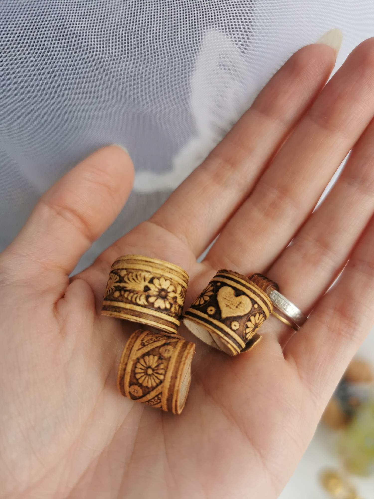 A close-up, top-down shot features a person's open hand holding three small, decorative cylindrical objects. These objects are made of light-colored wood or birch bark and are adorned with intricate pyrography (wood burning) designs in dark brown. One object in the upper left displays a repeating floral pattern. Another, positioned to the right, features a central heart design with cross-hatching, flanked by smaller floral motifs. The third object, located at the bottom, showcases a prominent flower design surrounded by patterned bands.
The hand has slender fingers and well-maintained nails. On the ring finger, two rings are visible: a silver-toned band with a subtle etched pattern and a plain gold-toned band stacked below it. The lines and texture of the palm are clearly visible.
The background is a soft, diffused, light-colored fabric, likely a sheer curtain or screen, showing a distinct grid pattern from its weave. This indicates an indoor setting, illuminated by natural, diffused daylight. In the far bottom right, out-of-focus elements, possibly other decorative items or beads in gold and green hues, are faintly discernible. The image focuses on presenting the craftsmanship of the wooden items. No visible text is present.